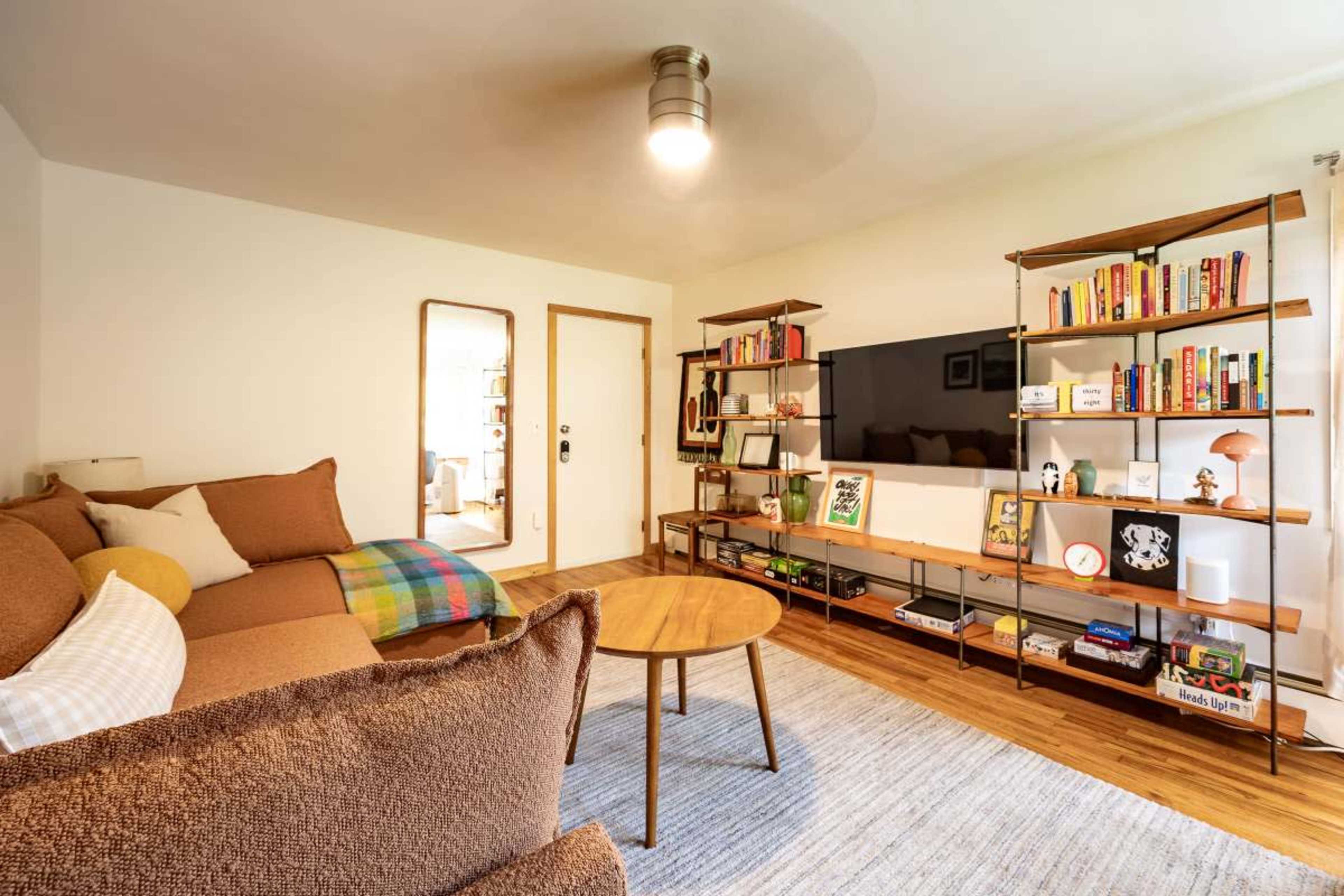 The image shows a cozy living room with a brown sectional sofa, a circular wooden coffee table, and a shelving unit holding books and decorative items, while a television is mounted on the wall opposite the sofa.