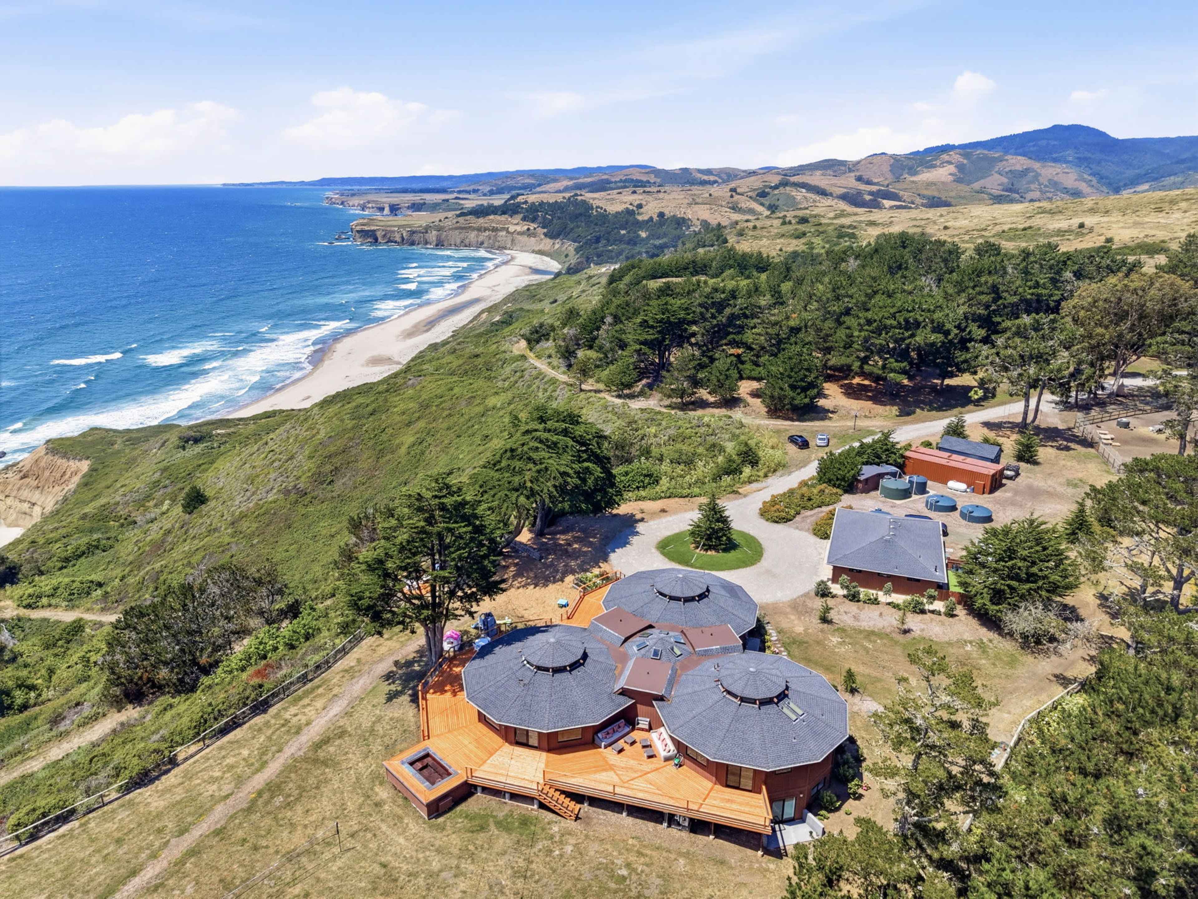 The image shows a coastal property with several round buildings surrounded by greenery and a view of the ocean and shoreline in the background.