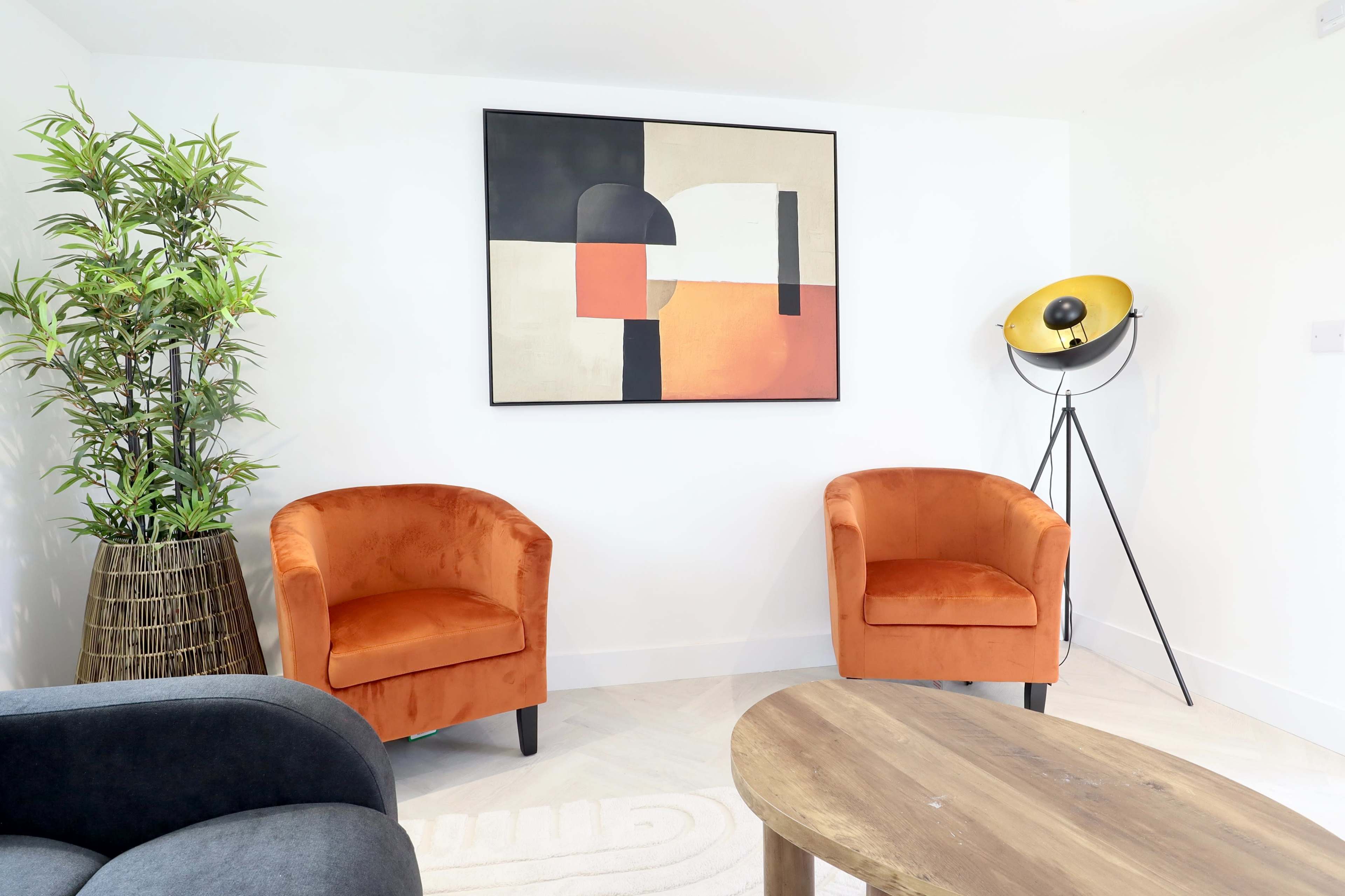 The scene features two orange armchairs flanking a modern abstract painting on a white wall, with a potted plant and a floor lamp in the corner.