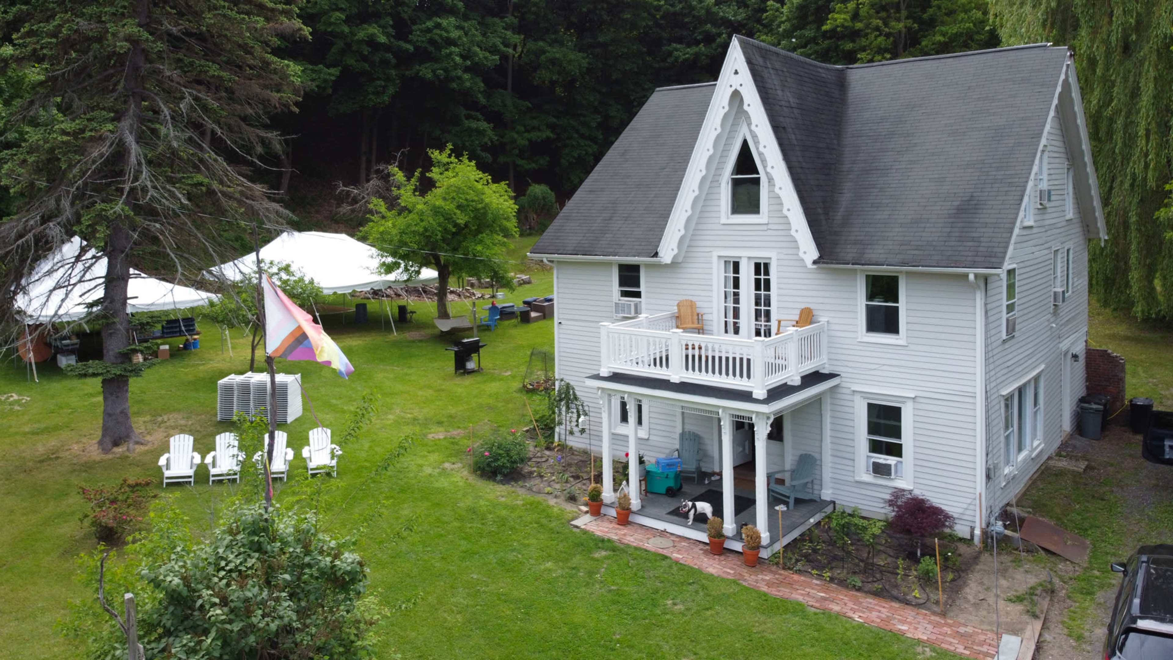 A two-story white house with a pitched roof has a porch and is surrounded by a well-kept lawn featuring chairs, tents, and trees.