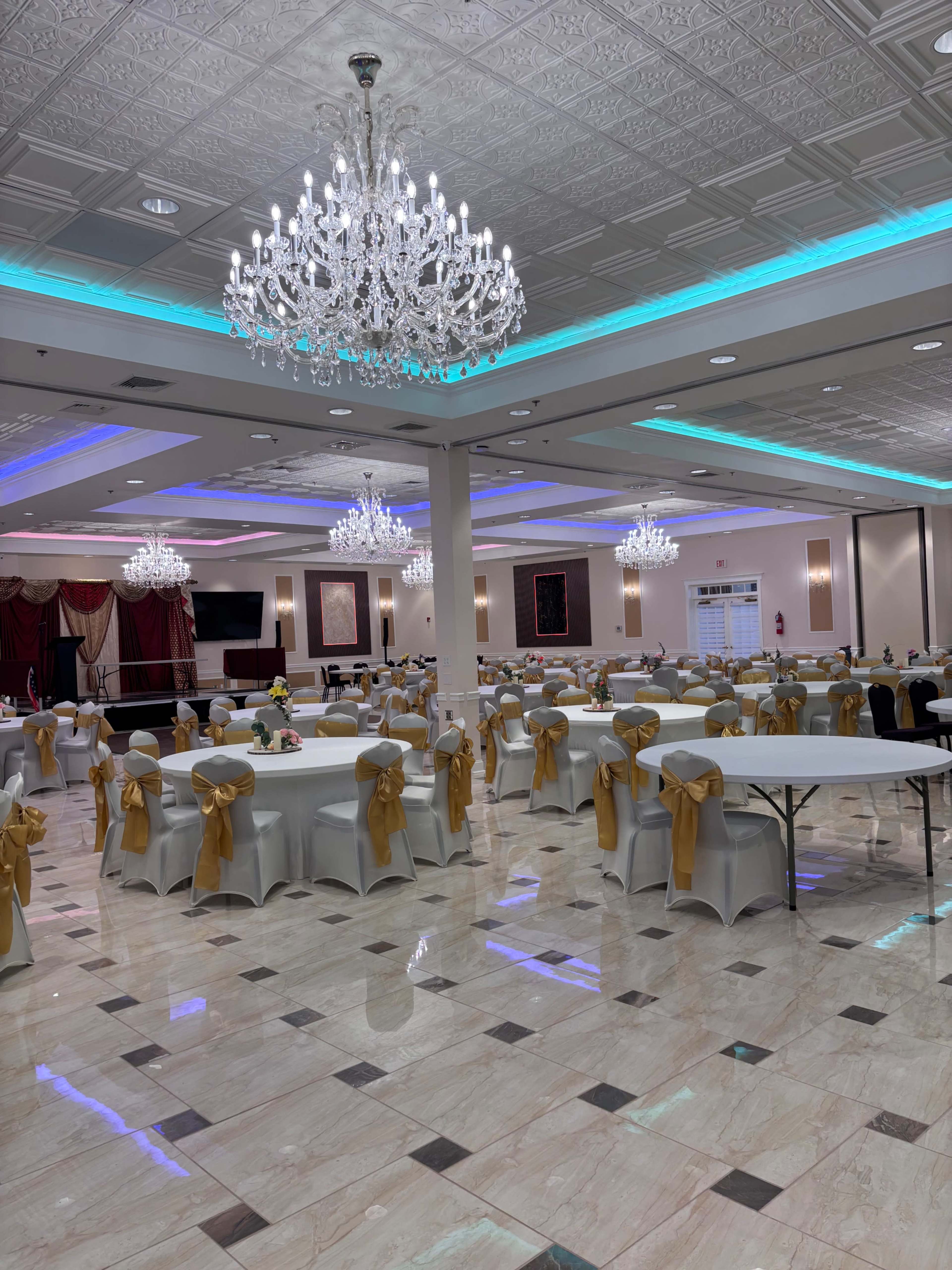 The image shows a large banquet hall with tables covered in white linens and decorated with yellow satin bows, illuminated by elegant chandeliers.
