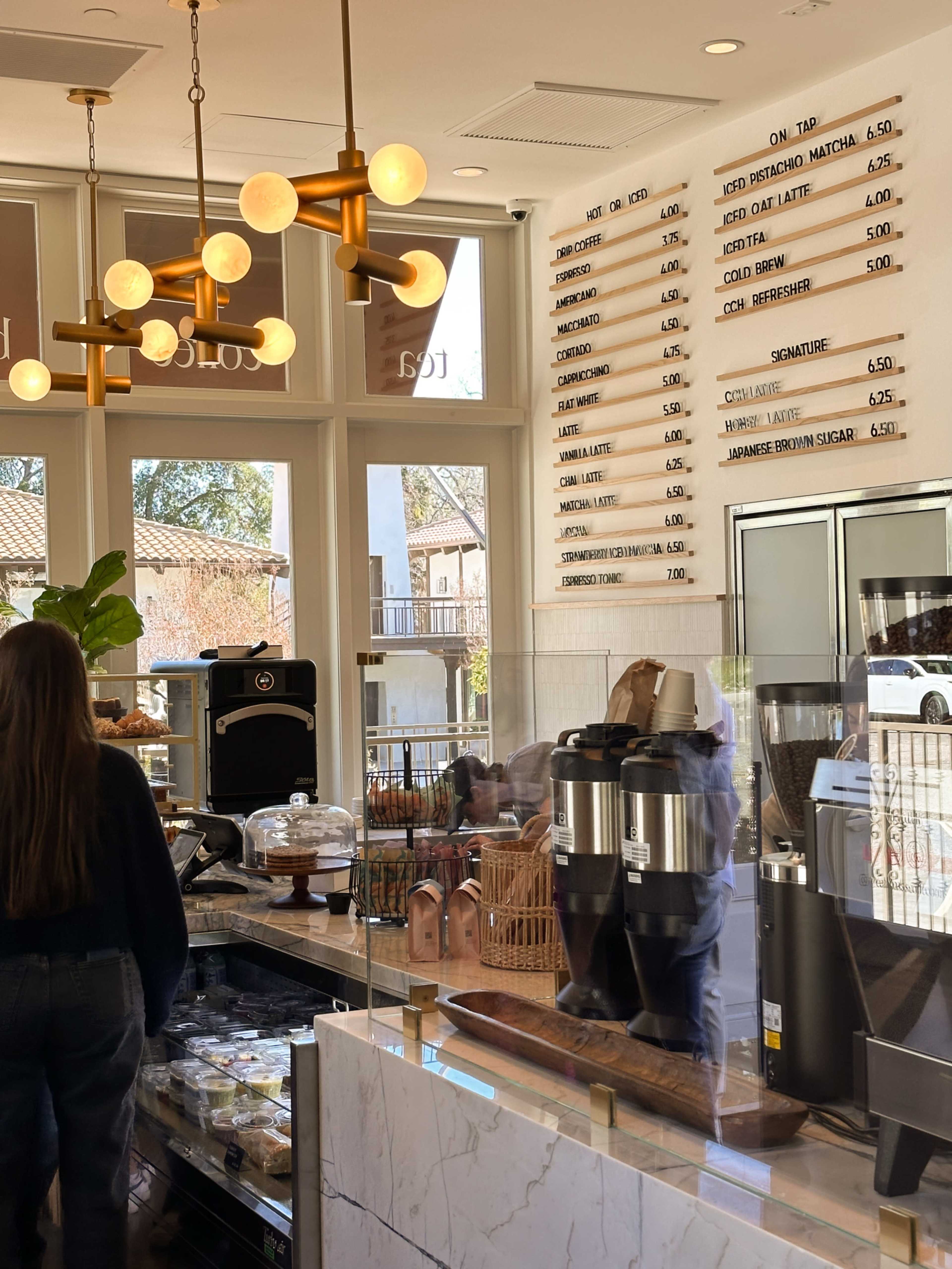 The image shows a cafe interior featuring a menu board with various drink options and prices, alongside a glass display case with baked goods.