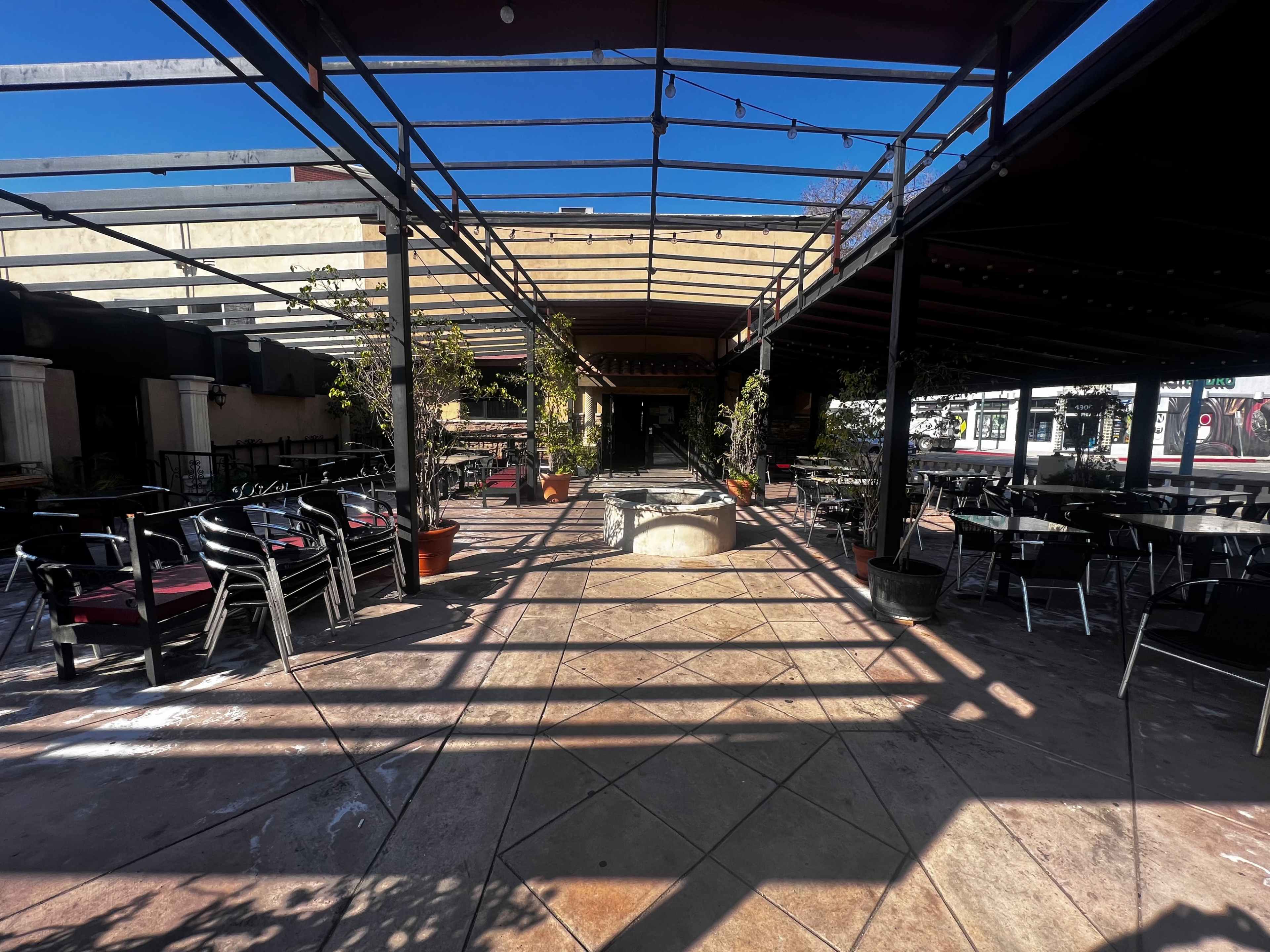 An empty outdoor dining area with stacked chairs and tables under a canopy, surrounded by potted plants.