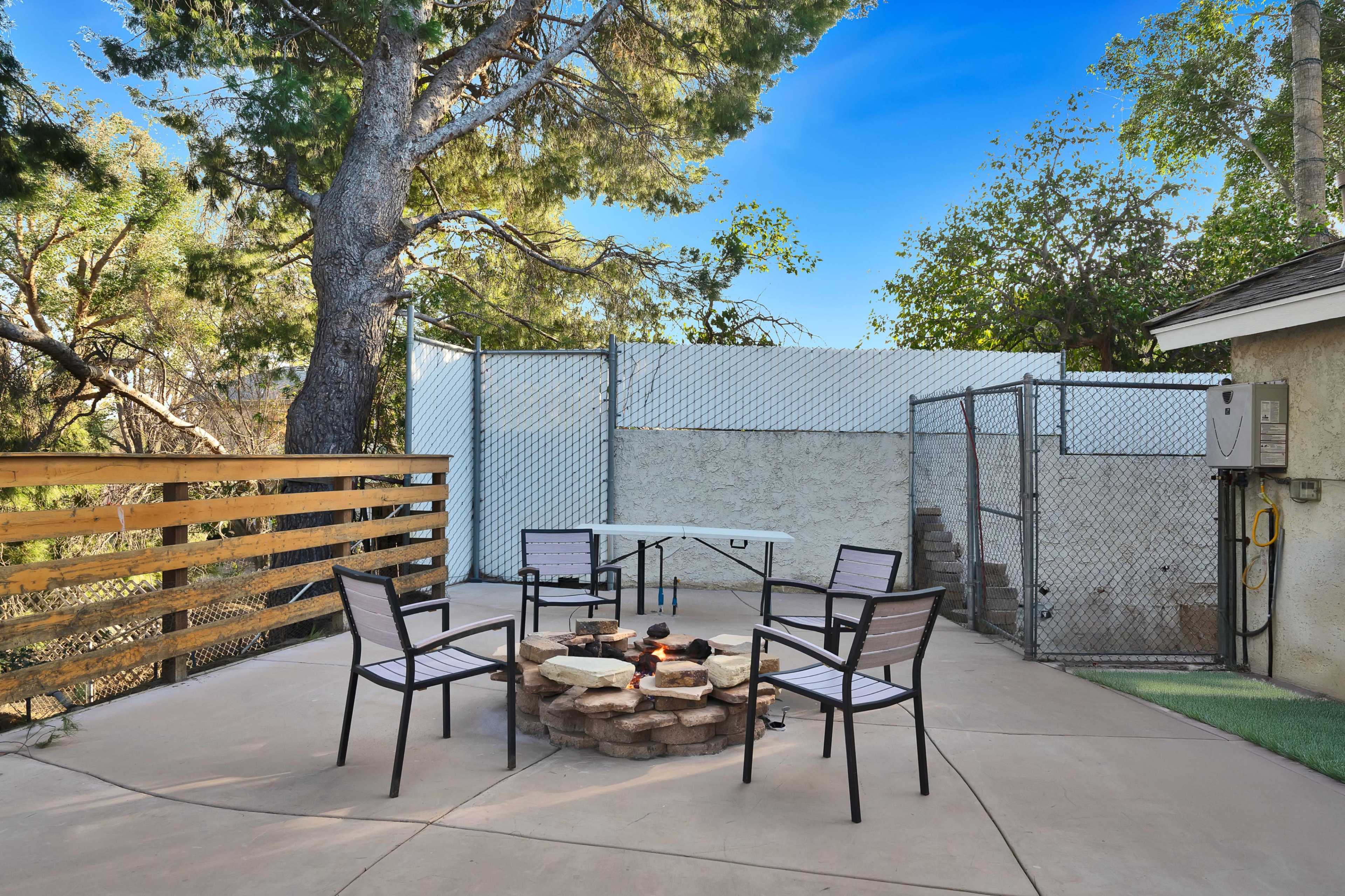 The scene shows a patio area with a circular stone fire pit surrounded by four chairs, adjacent to a fenced yard and trees.