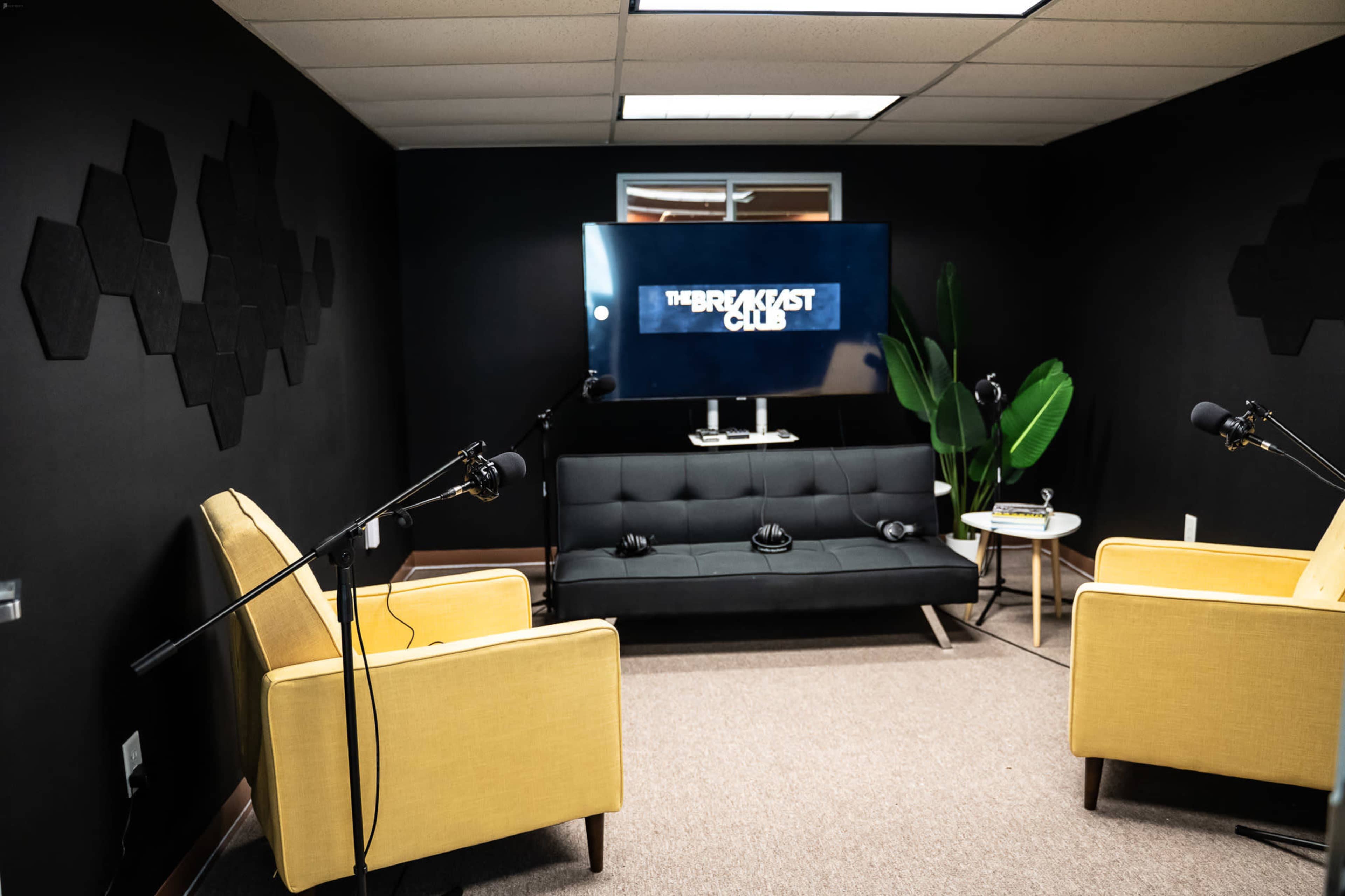 The image shows a podcast recording studio with two yellow chairs, a black couch, a television displaying "THE BREAKFAST CLUB," and microphones set up for recording.