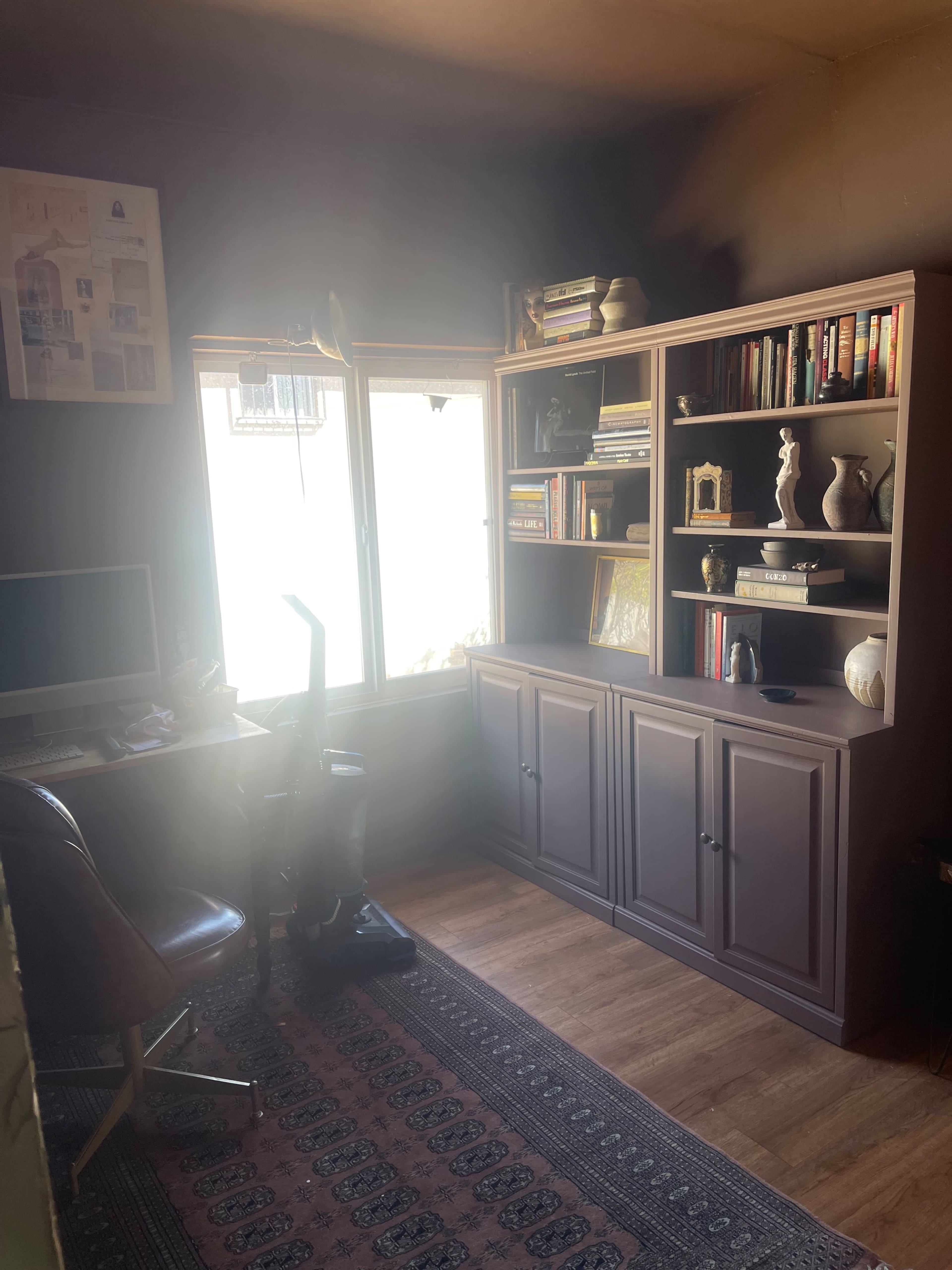 The image shows a small office space with a desk, an ergonomic chair, bookshelves filled with books and decorative items, and a window allowing natural light into the room.