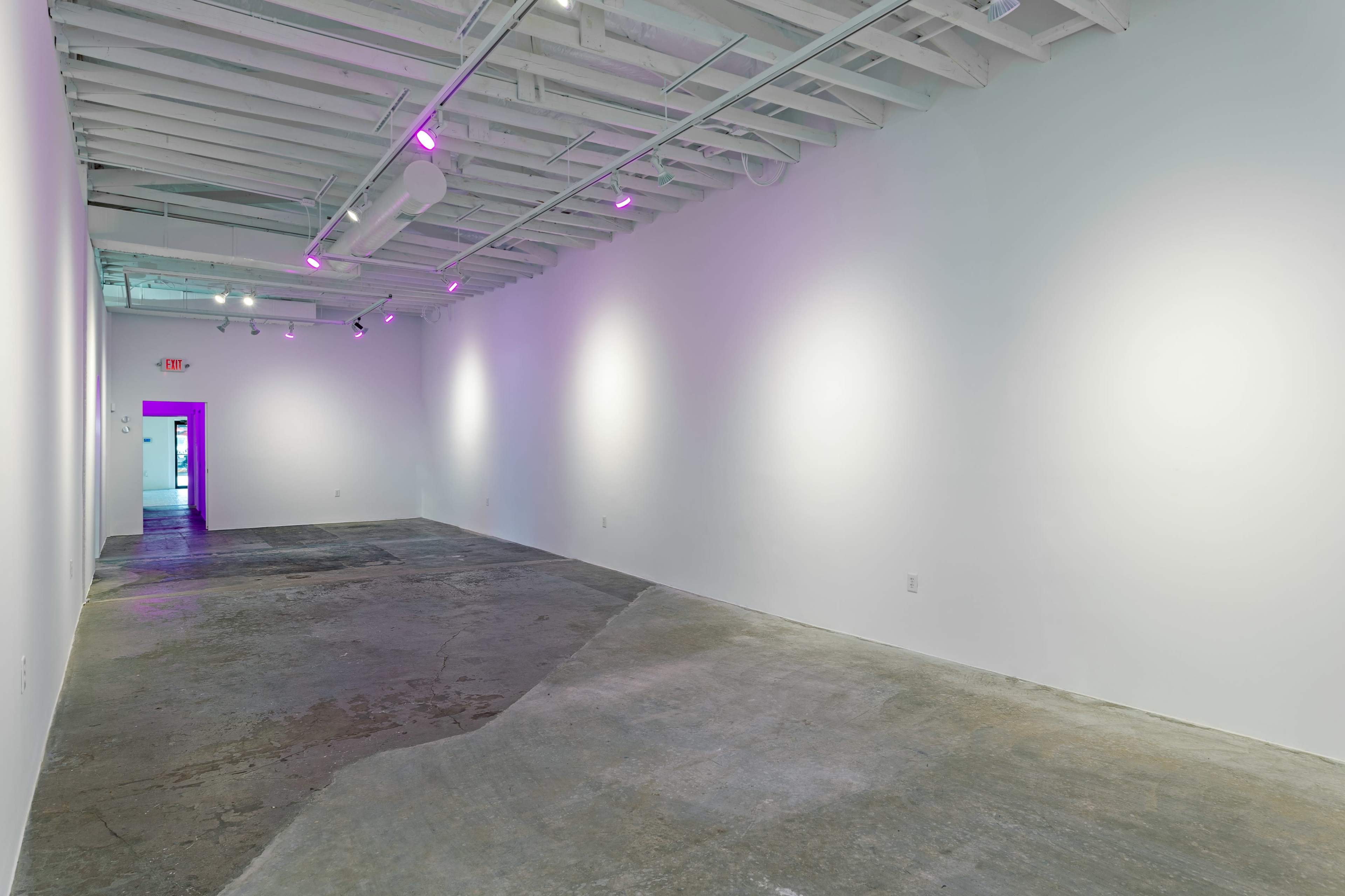 A spacious, minimalist art gallery features a concrete floor and white walls, with purple lighting illuminating the entrance at the far end.