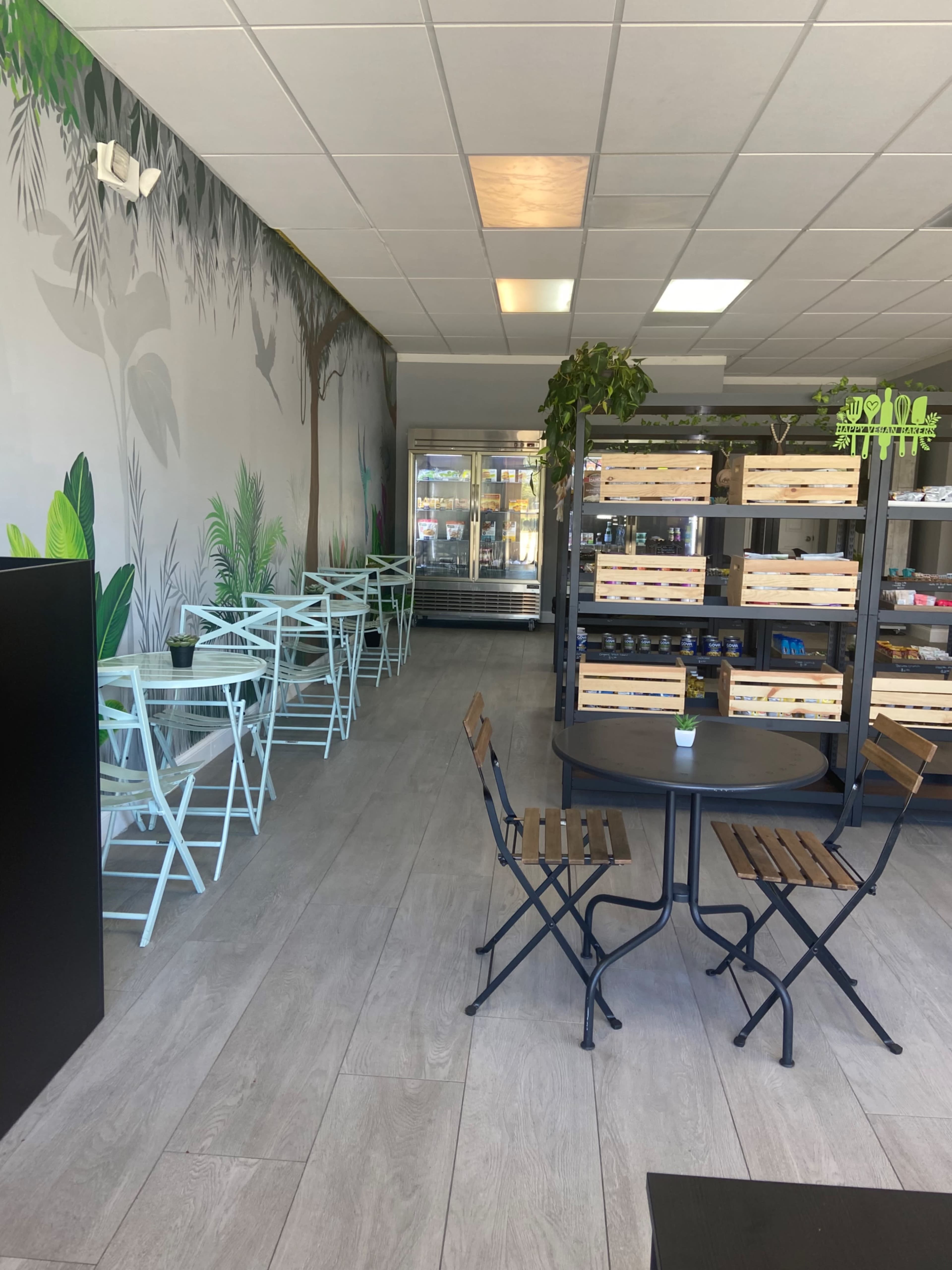 The image shows a modern café interior with light blue folding chairs and a dark metal table, surrounded by a mural of greenery and shelving displaying goods.