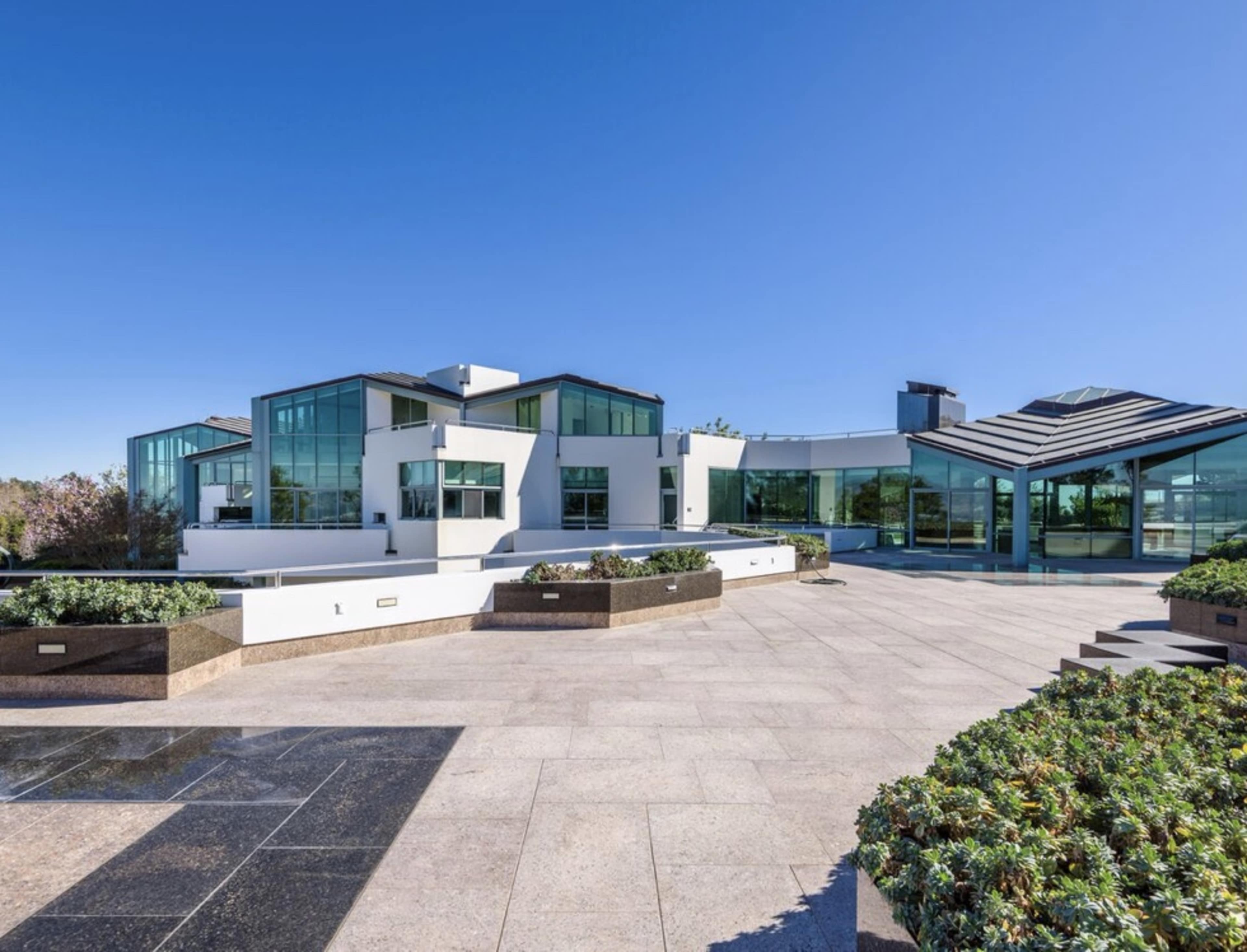 A modern two-story house featuring large glass windows and a flat roof, surrounded by landscaped gardens and stone pathways.
