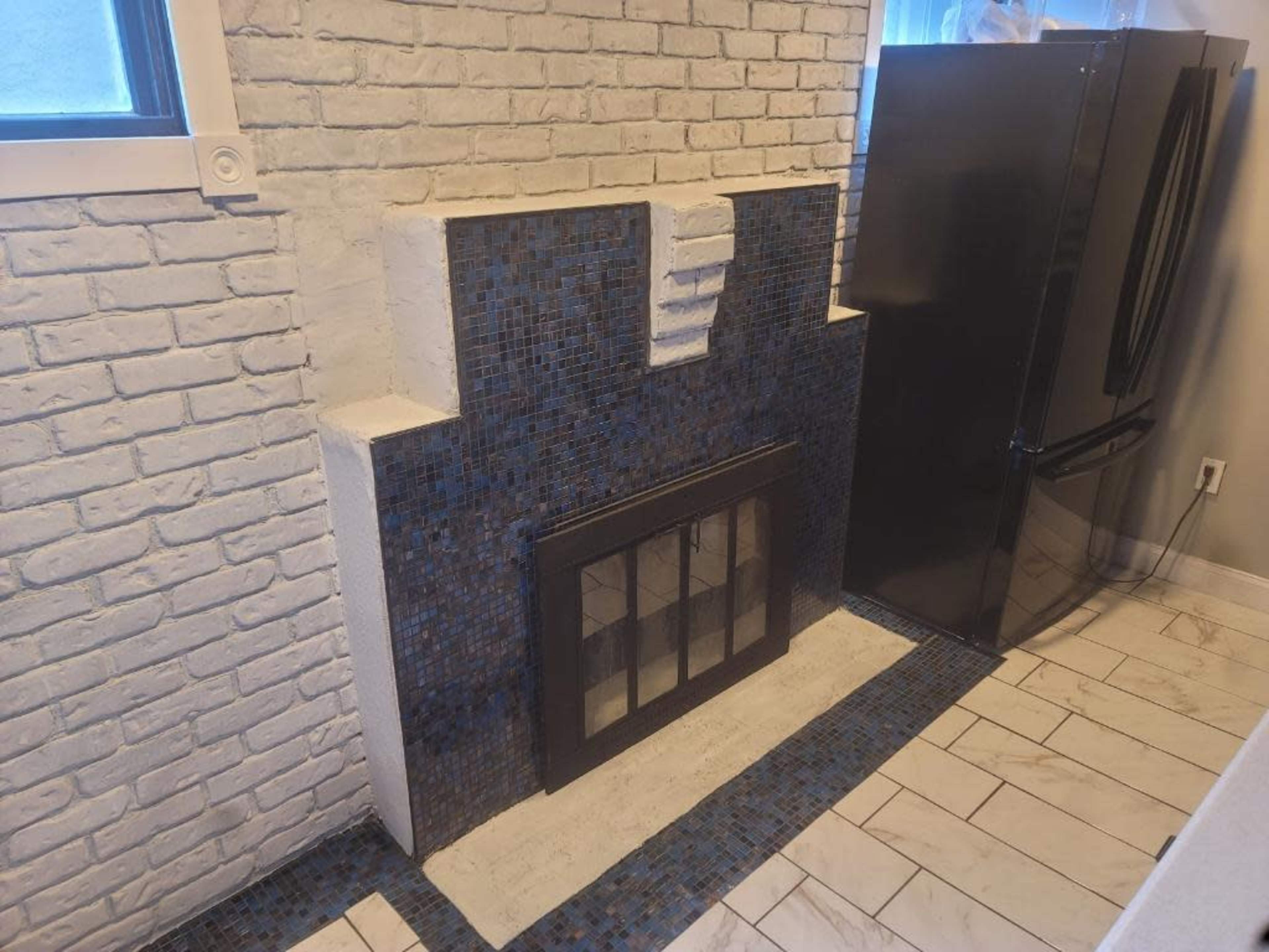 A white brick wall with a tiled fireplace and a black refrigerator beside it in a kitchen setting.