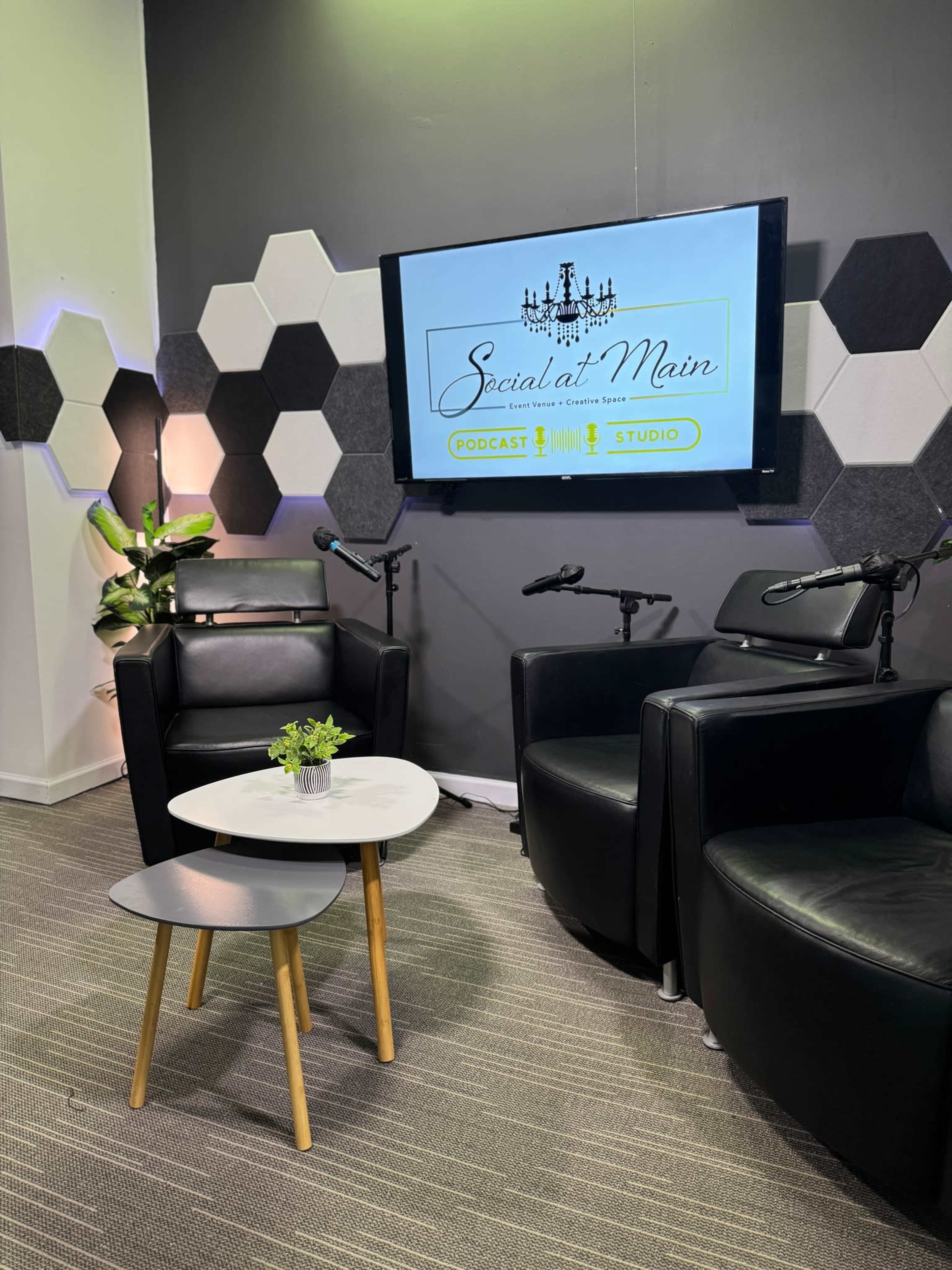 The image shows a podcast studio with two black armchairs, a small white table, and a display screen showcasing the "Social at Main" logo on a gray wall decorated with hexagonal panels.