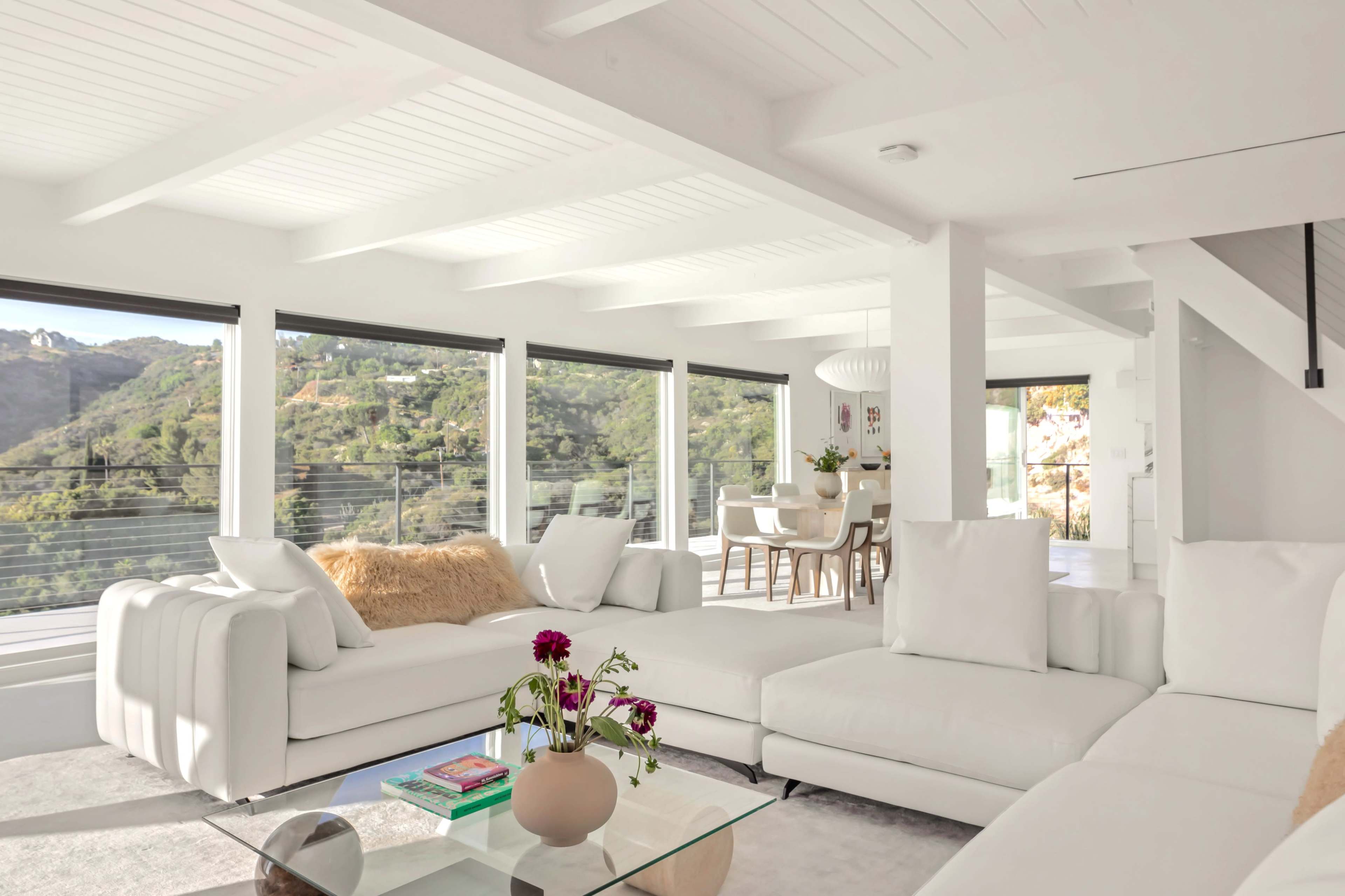A bright, modern living room with white furniture, large windows offering a view of the green hills outside, and a dining area in the background.