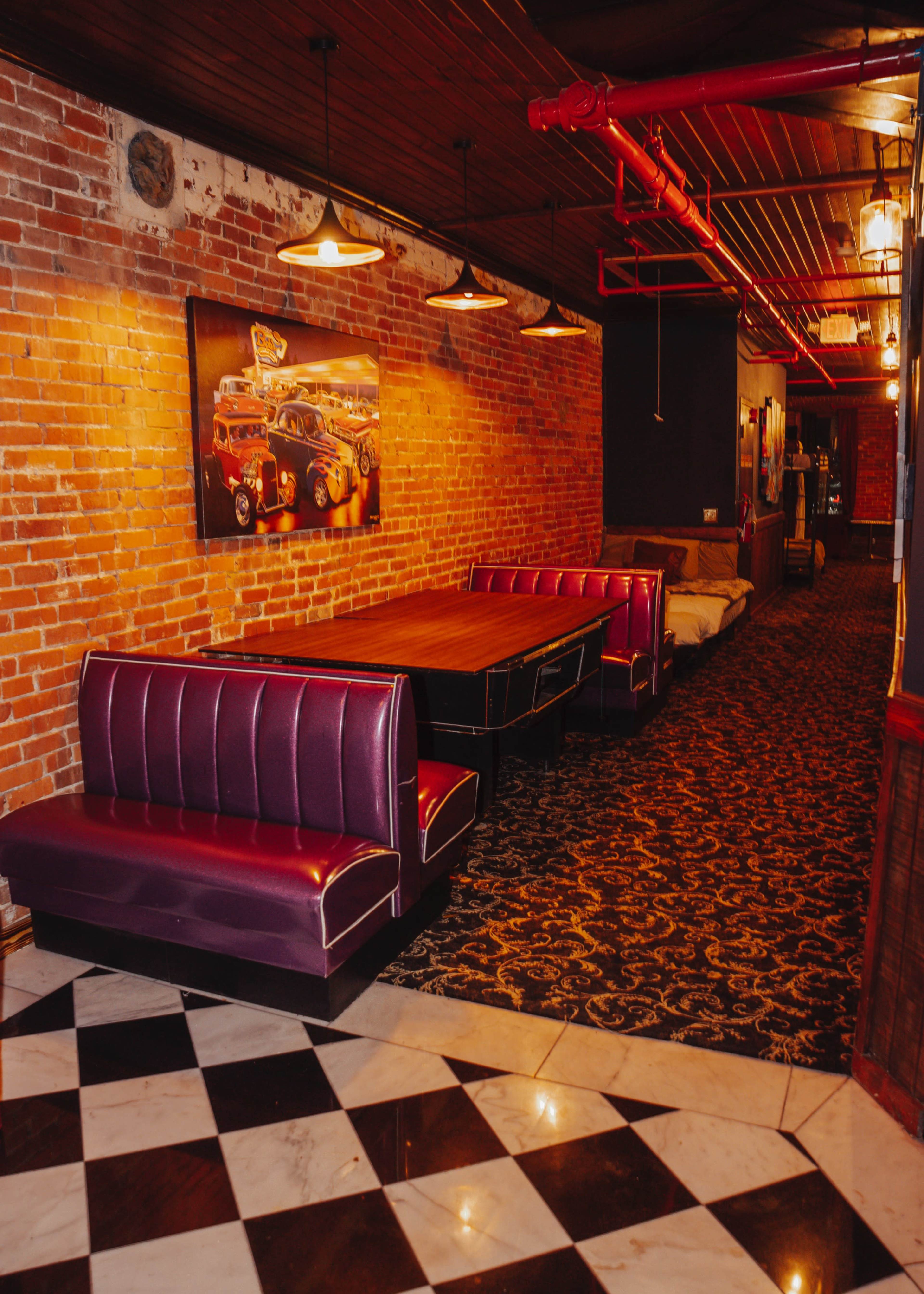 The image shows a dimly lit interior space with brick walls, featuring a long table surrounded by purple booth seating and a checkered floor.
