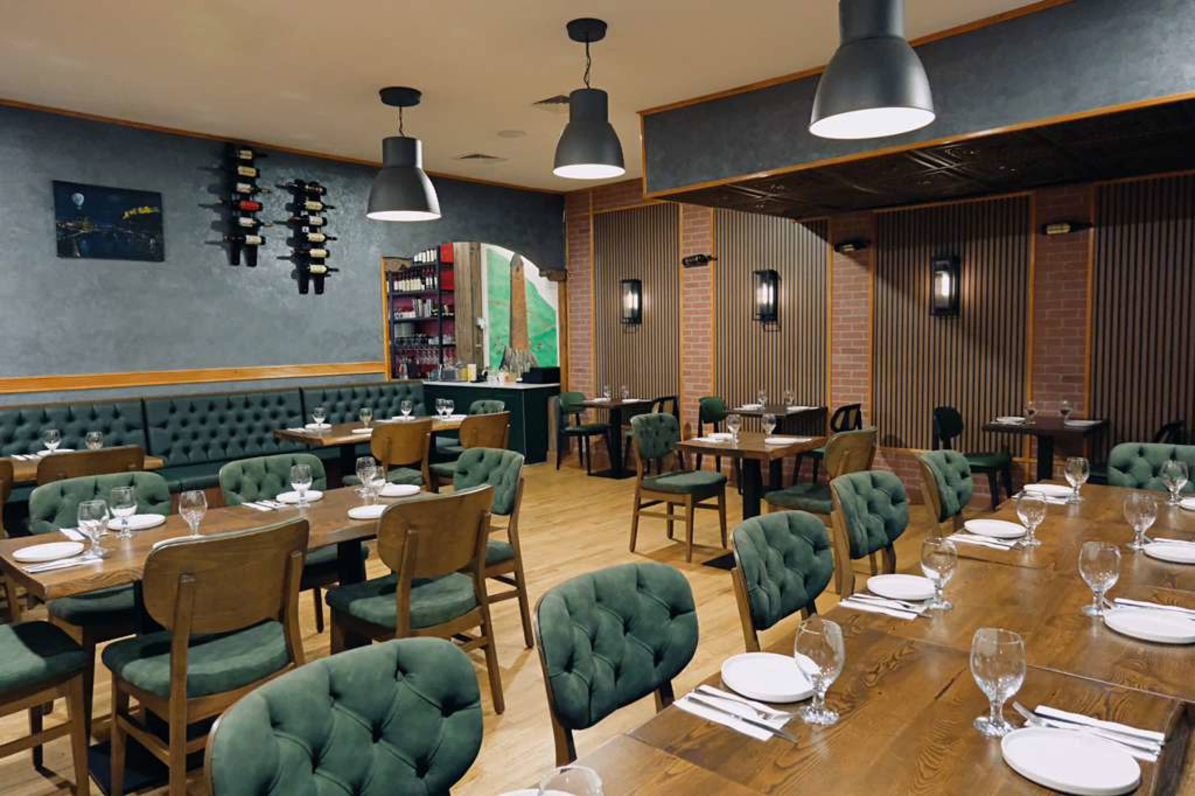 The image shows the interior of a restaurant featuring wooden tables and green upholstered chairs, with decorative lighting and wine storage displayed on the walls.