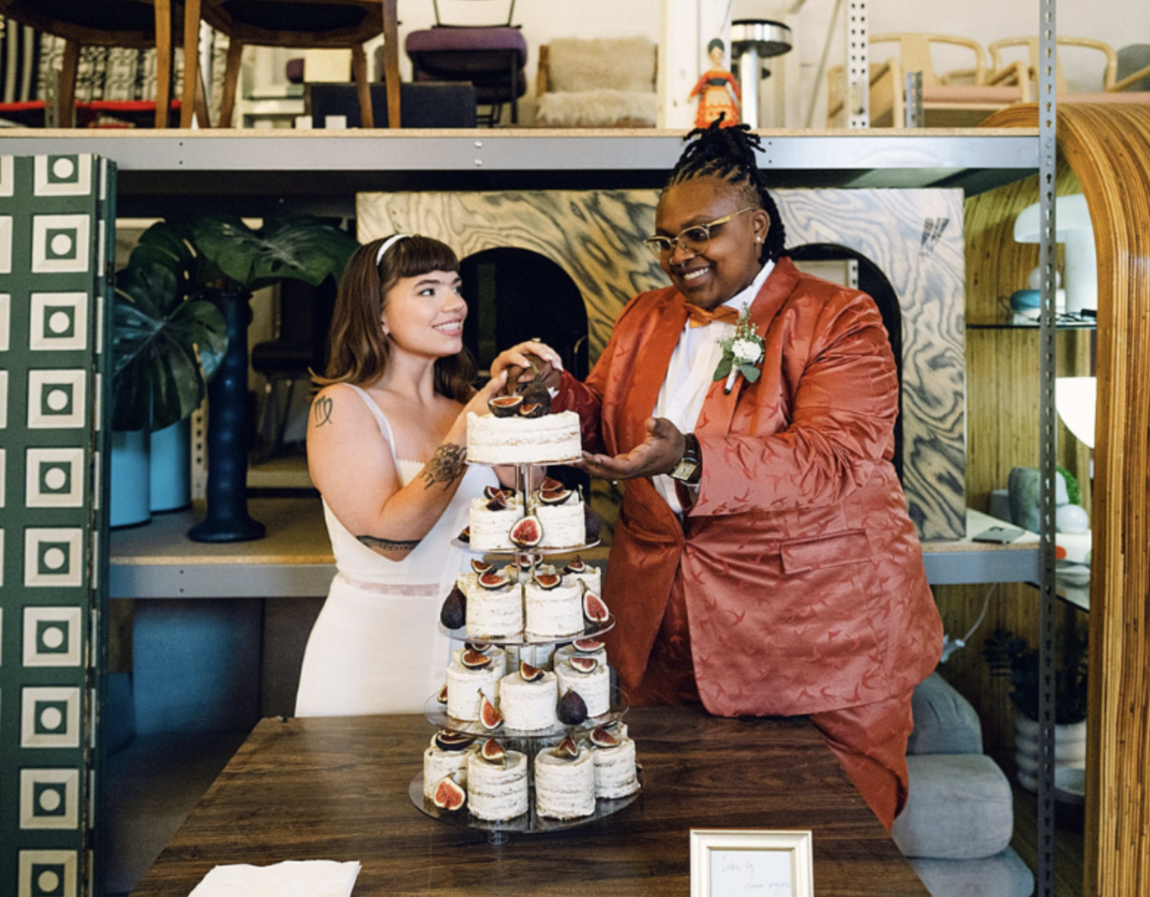 Two individuals stand beside a multi-tiered cake in a modern furniture setting, with one person playfully offering a piece of cake to the other.