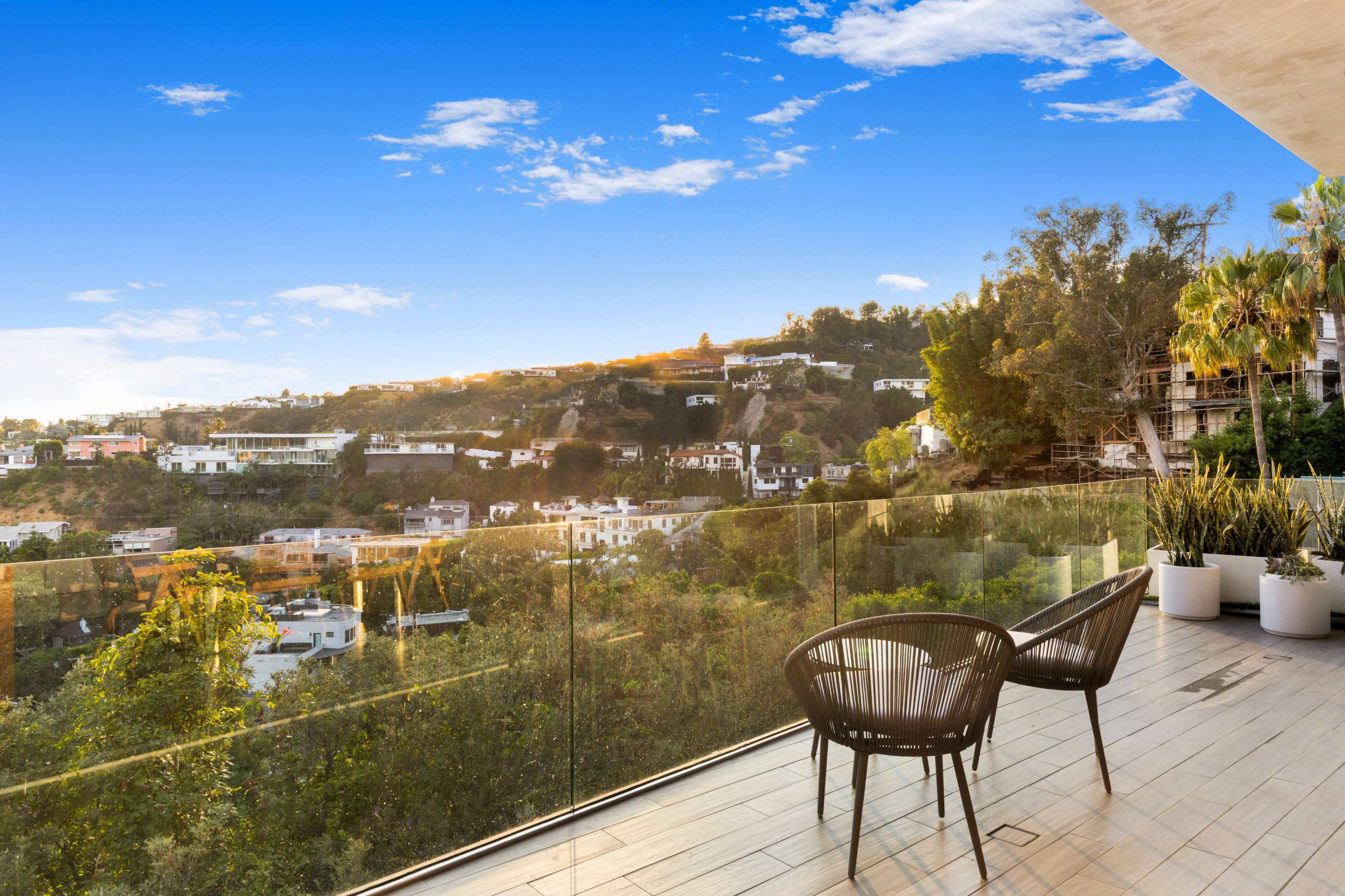 A balcony features two black chairs overlooking a hillside populated with white houses and greenery under a bright blue sky.