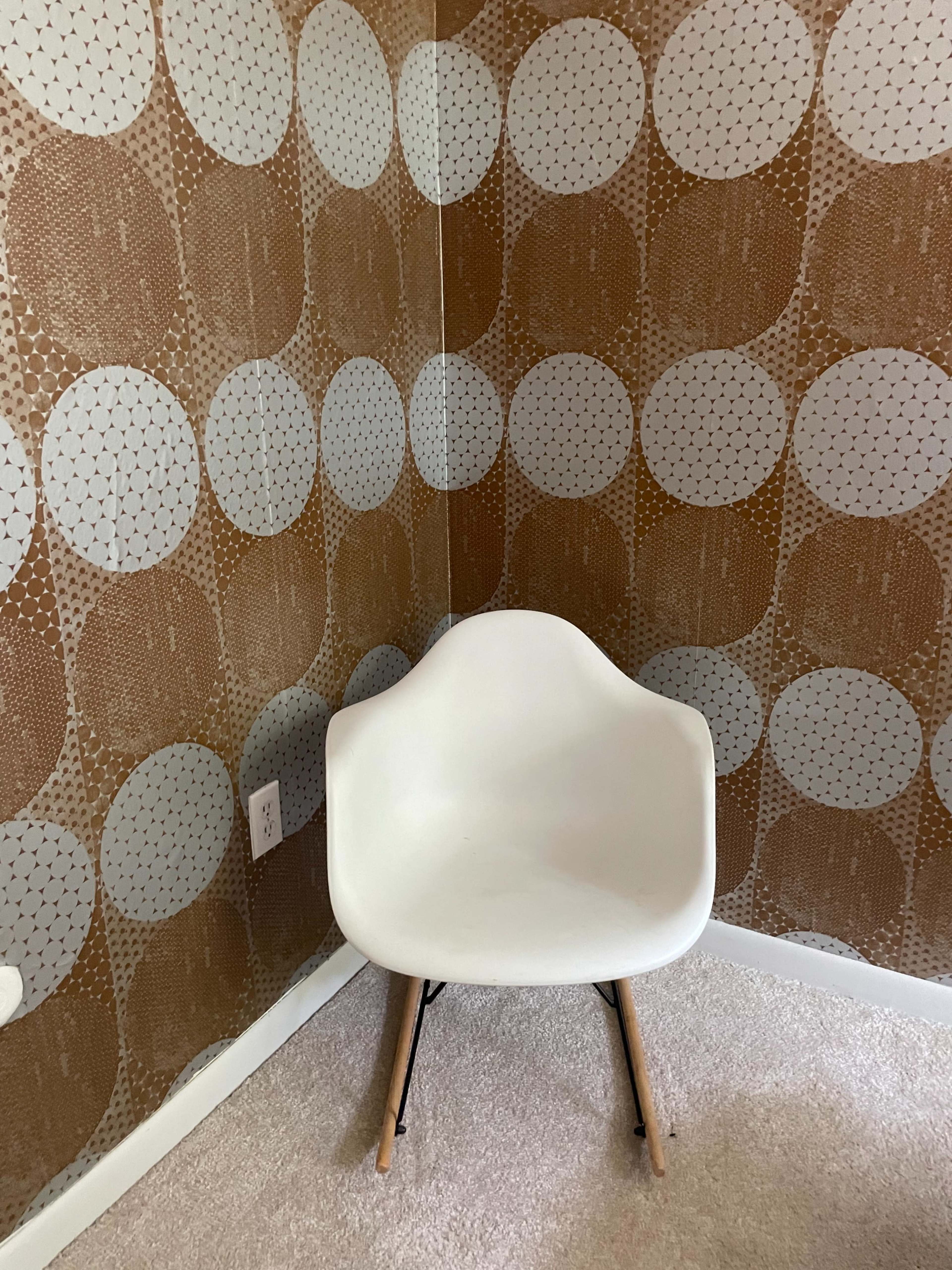 A white chair with wooden legs positioned in a corner beside patterned brown wallpaper featuring large white circles.