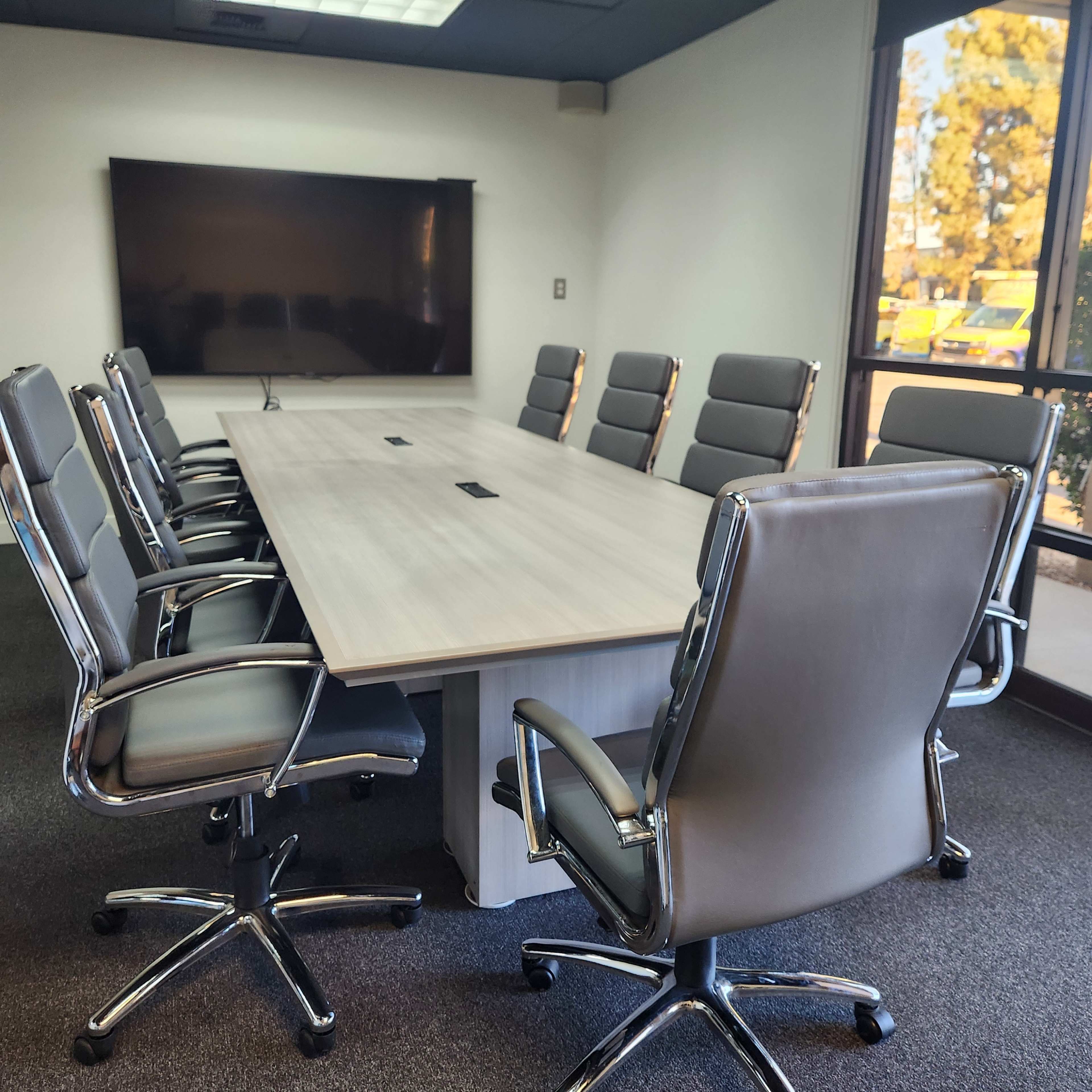 The image shows a modern conference room with a long wooden table surrounded by ergonomic chairs and a large screen mounted on the wall.