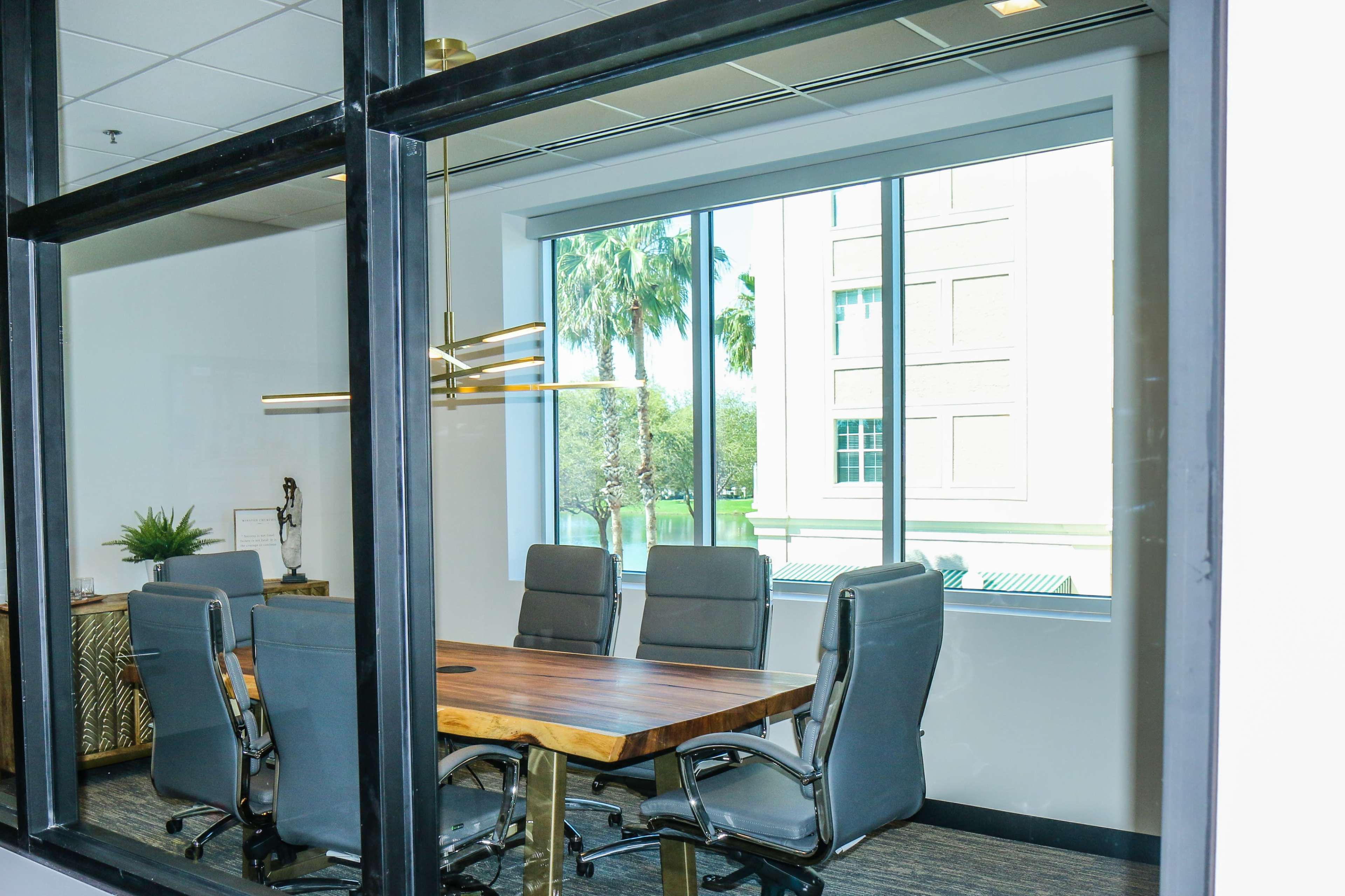 A conference room with a wood table and gray chairs is visible through a large window, showcasing palm trees outside.
