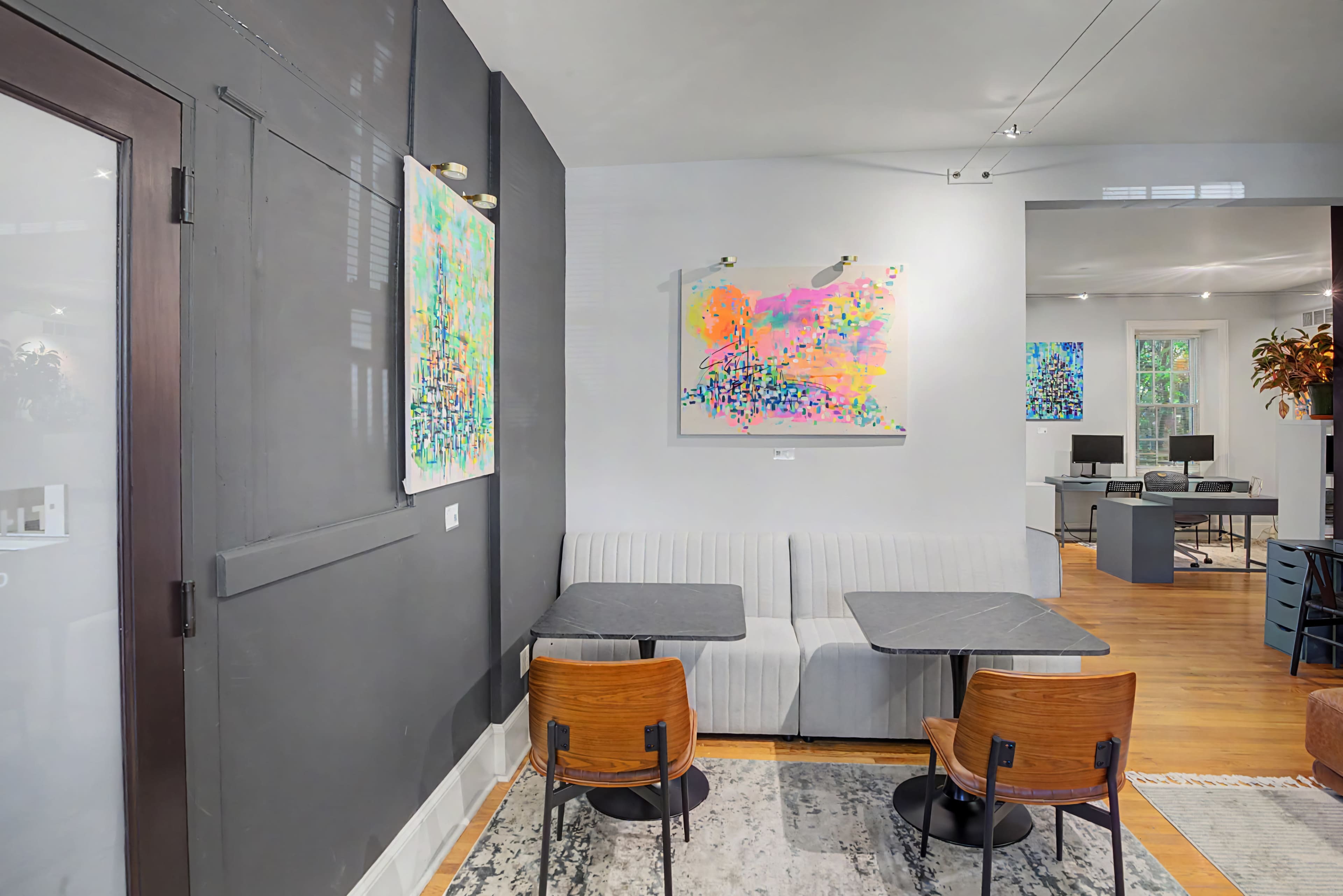 The image shows a modern coffee shop interior featuring two wooden chairs at a gray table, a gray banquette, and colorful abstract art on the walls.