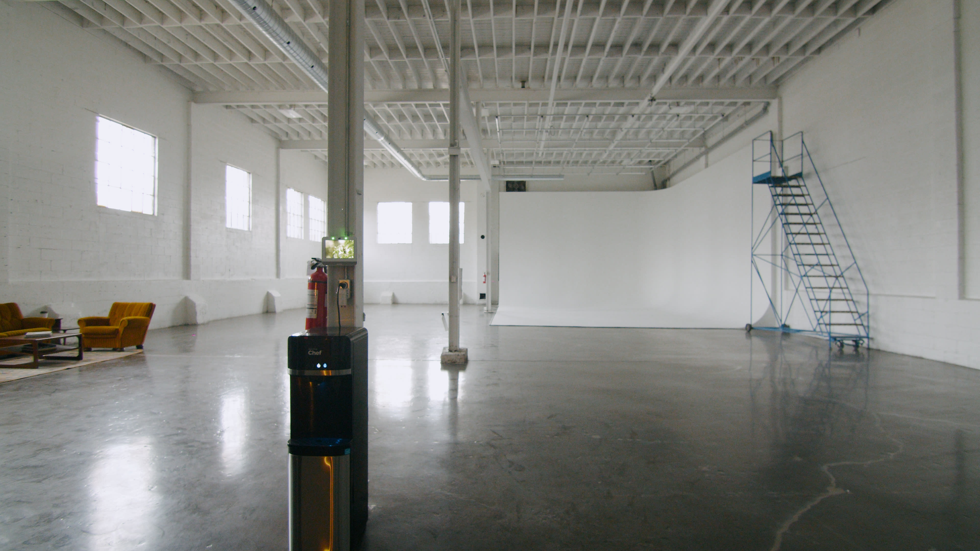 A spacious, minimalist studio with a polished concrete floor, large windows, and a blank white wall, along with a fire extinguisher and a ladder.