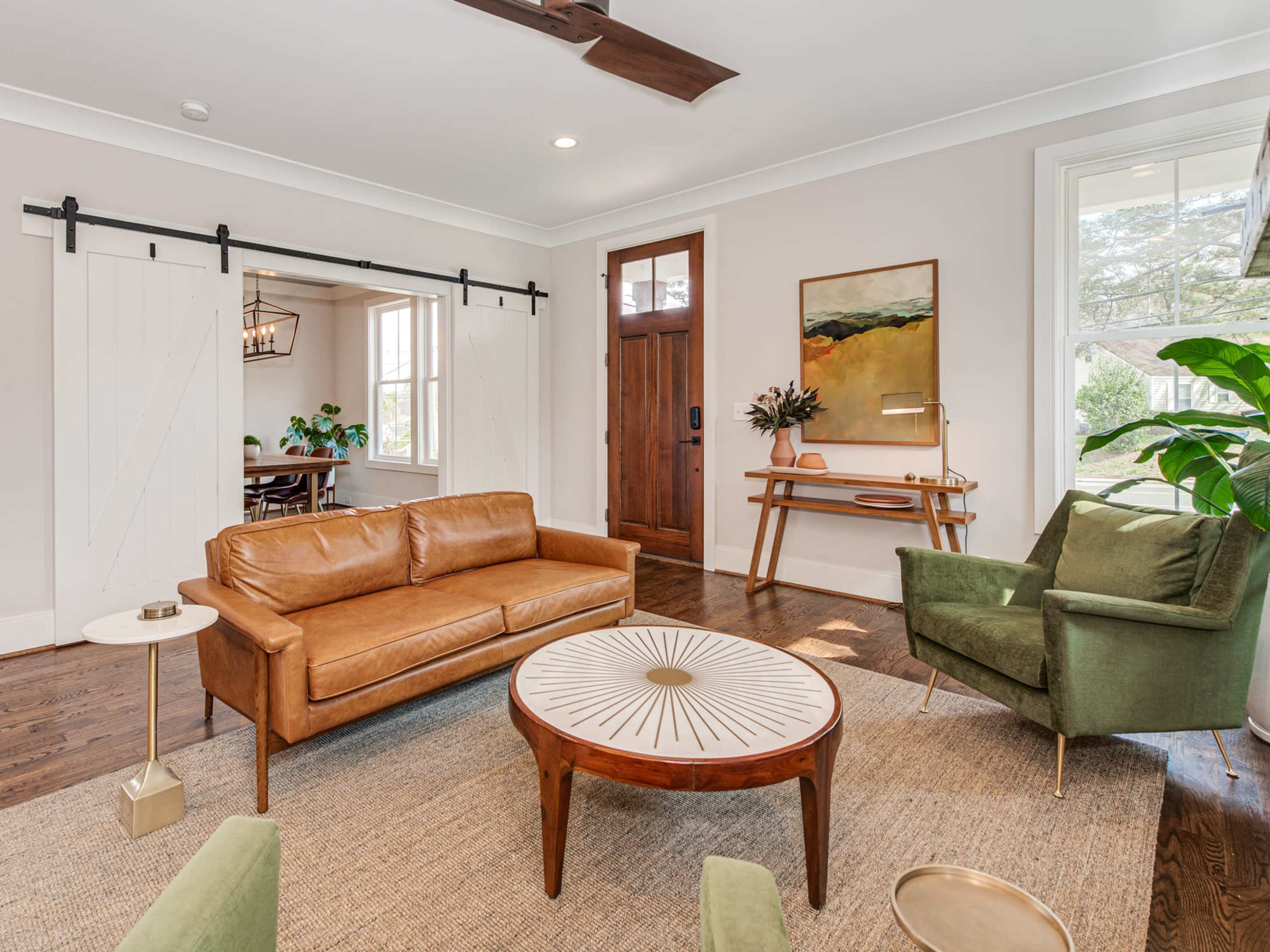 A cozy living room with a brown leather sofa, a green armchair, a round table, and a large artwork on the wall, along with a doorway leading to a dining area.