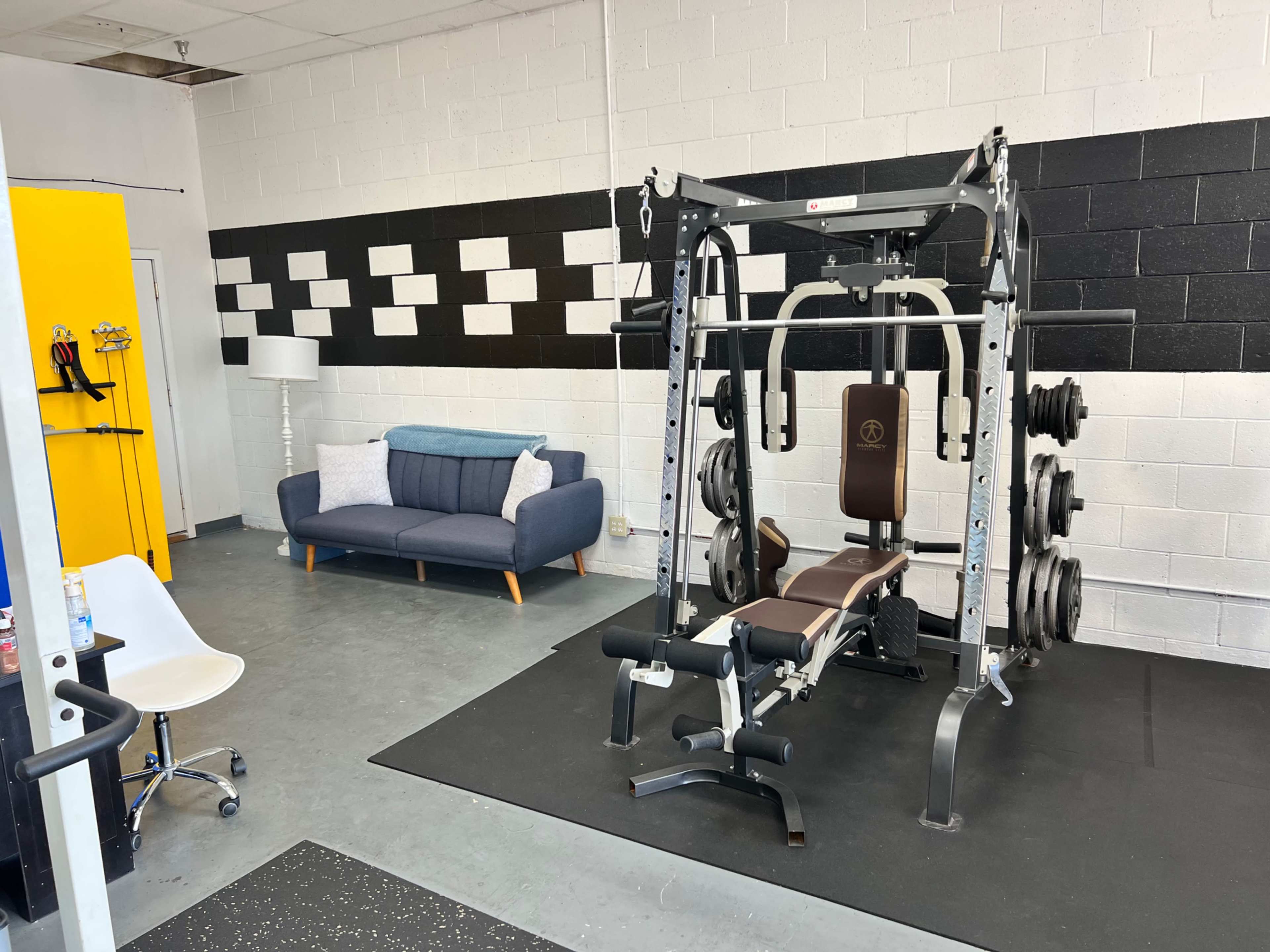 A gym space featuring a multi-station weight machine, a bench, a set of weights, a blue couch, and a yellow storage cabinet against a black and white accent wall.