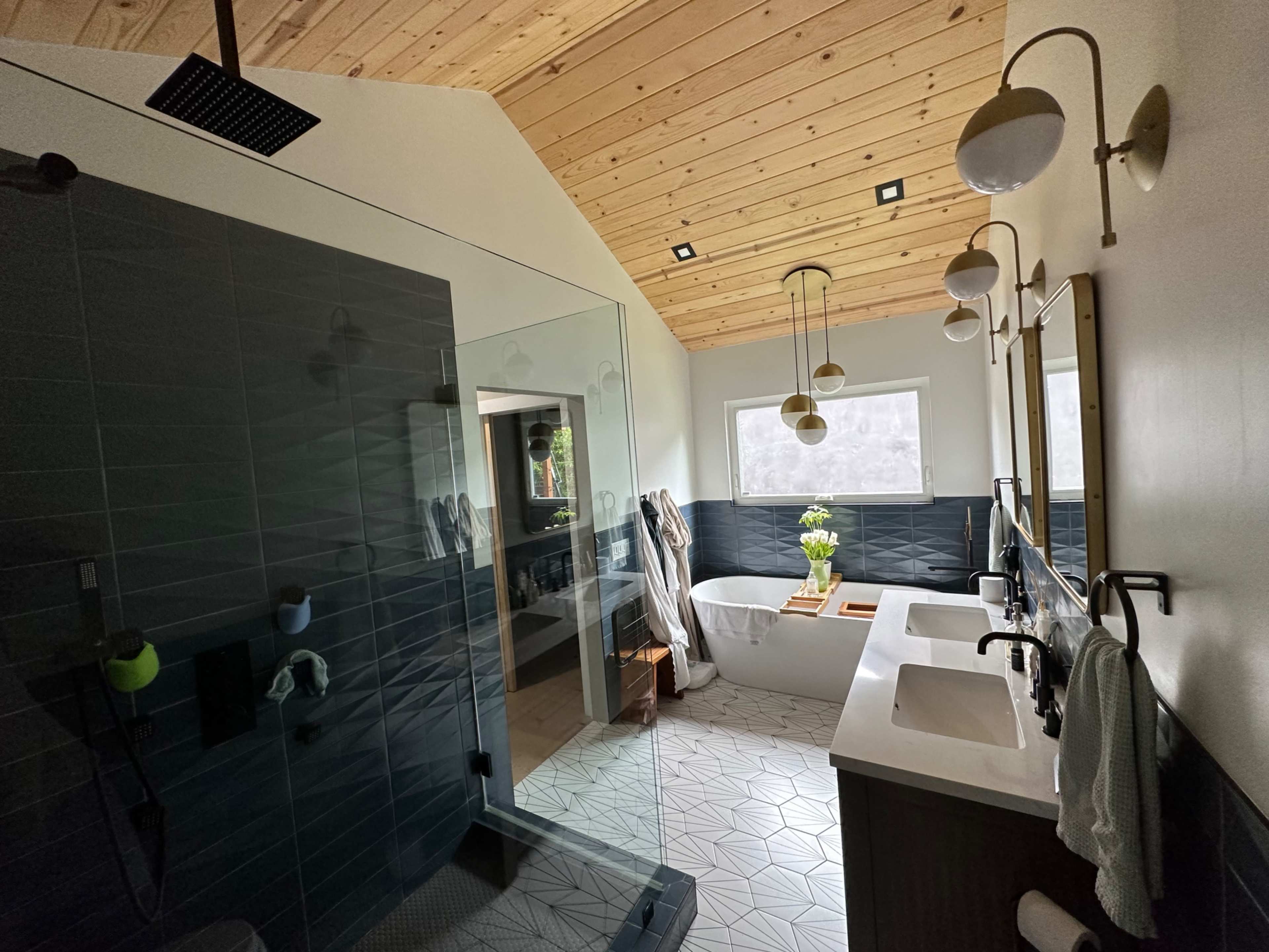 The image shows a modern bathroom featuring a glass shower, a freestanding bathtub, two sinks with mirrors, and a wooden ceiling.