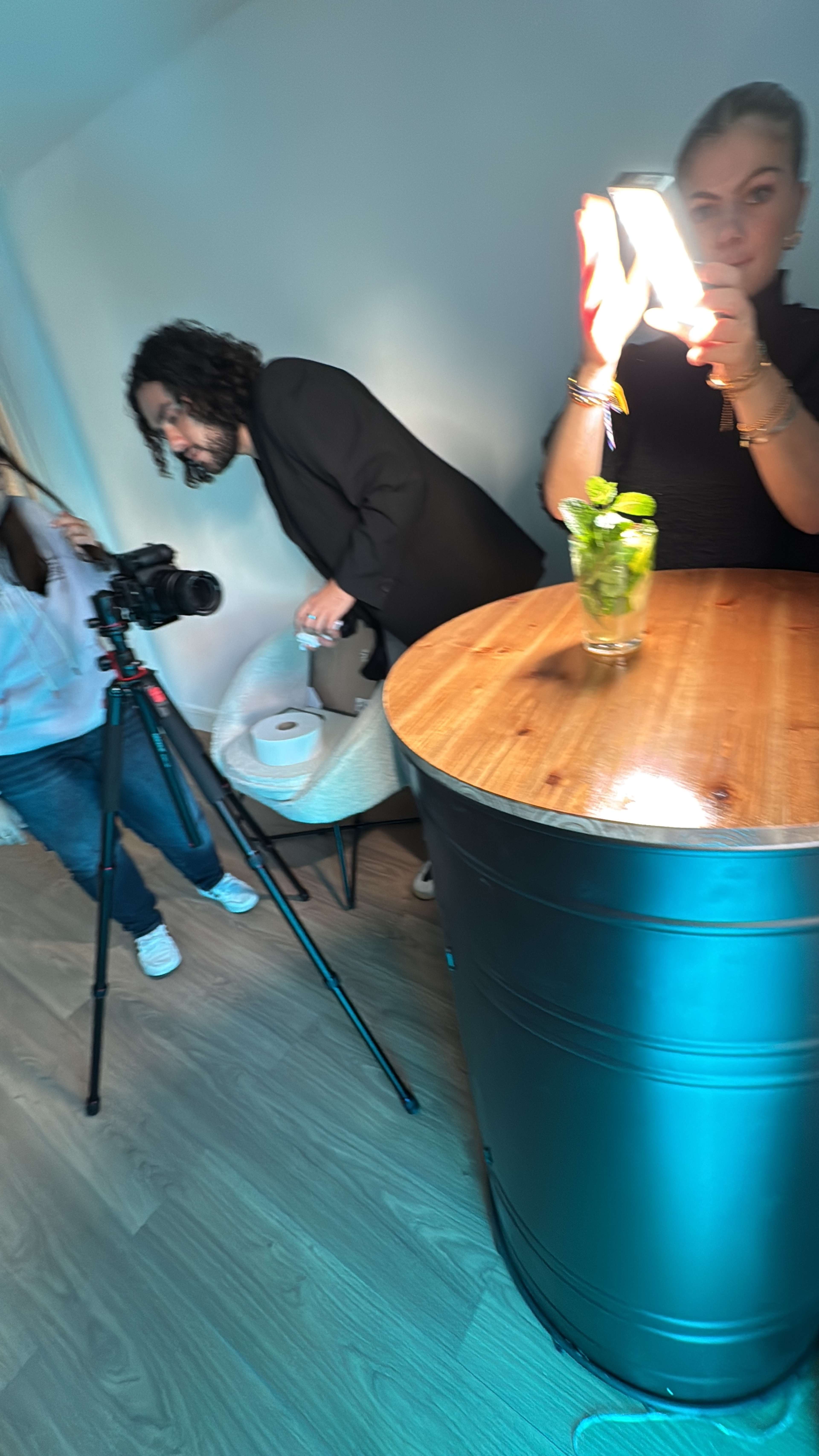 In a brightly lit room, a woman holds a light reflecting on a glass with a drink while a man in a black jacket adjusts equipment and another person sets up a camera nearby.