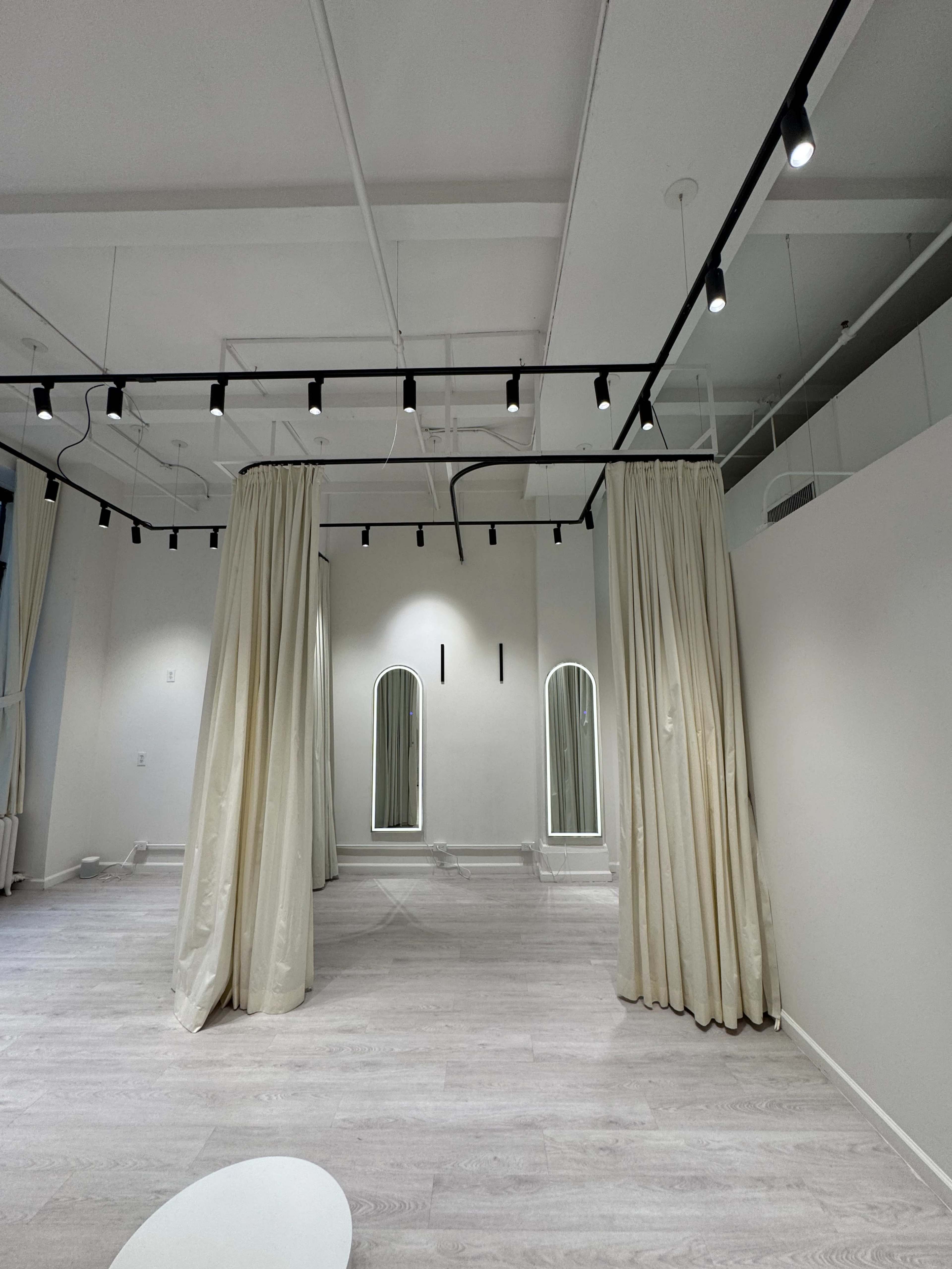 The image shows a spacious room with white walls, wooden flooring, and sheer curtains separating areas, complemented by three arch-shaped mirrors along one wall.