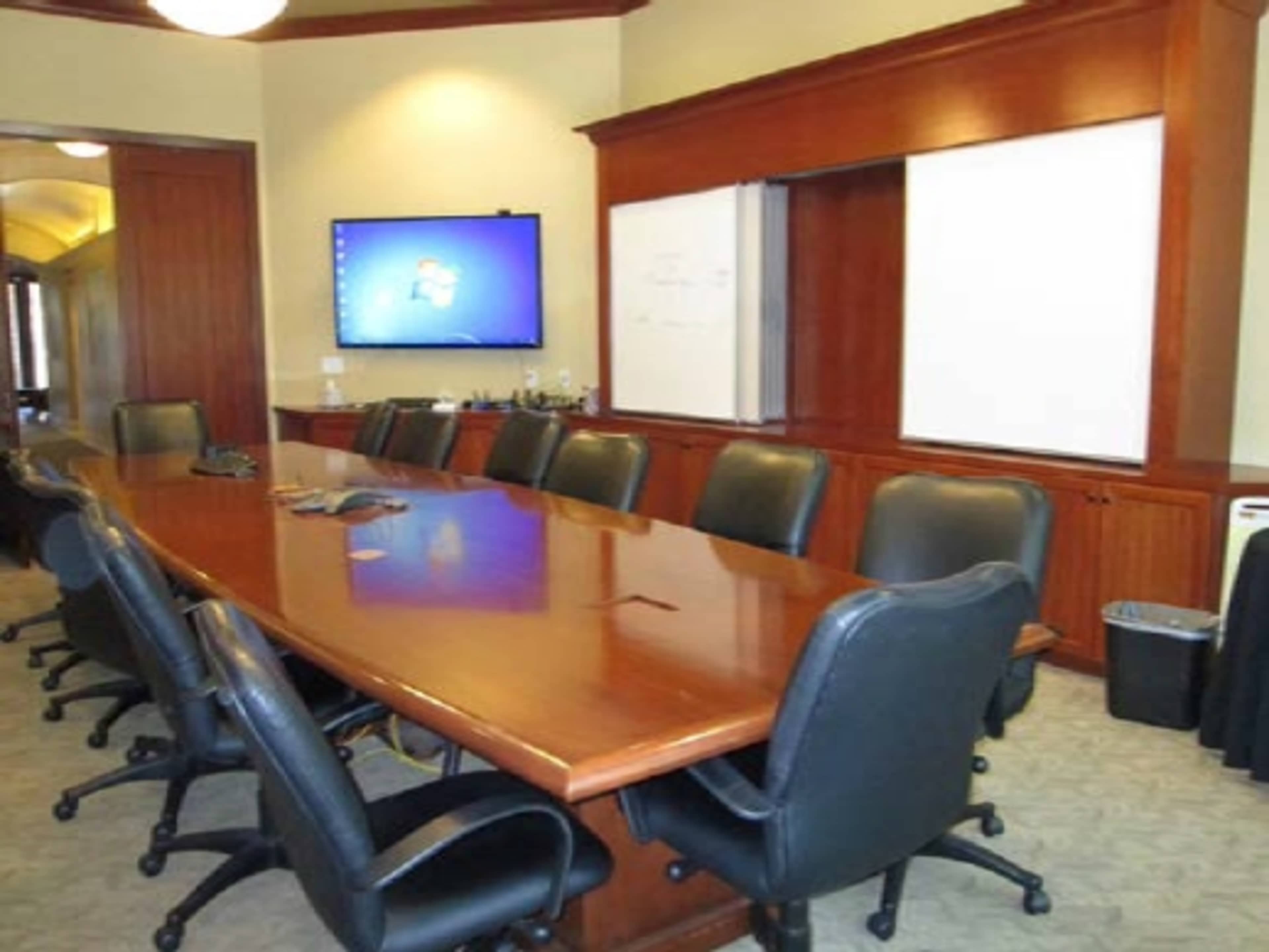 A large conference room features a long wooden table surrounded by leather chairs, with a screen and whiteboard on one wall.