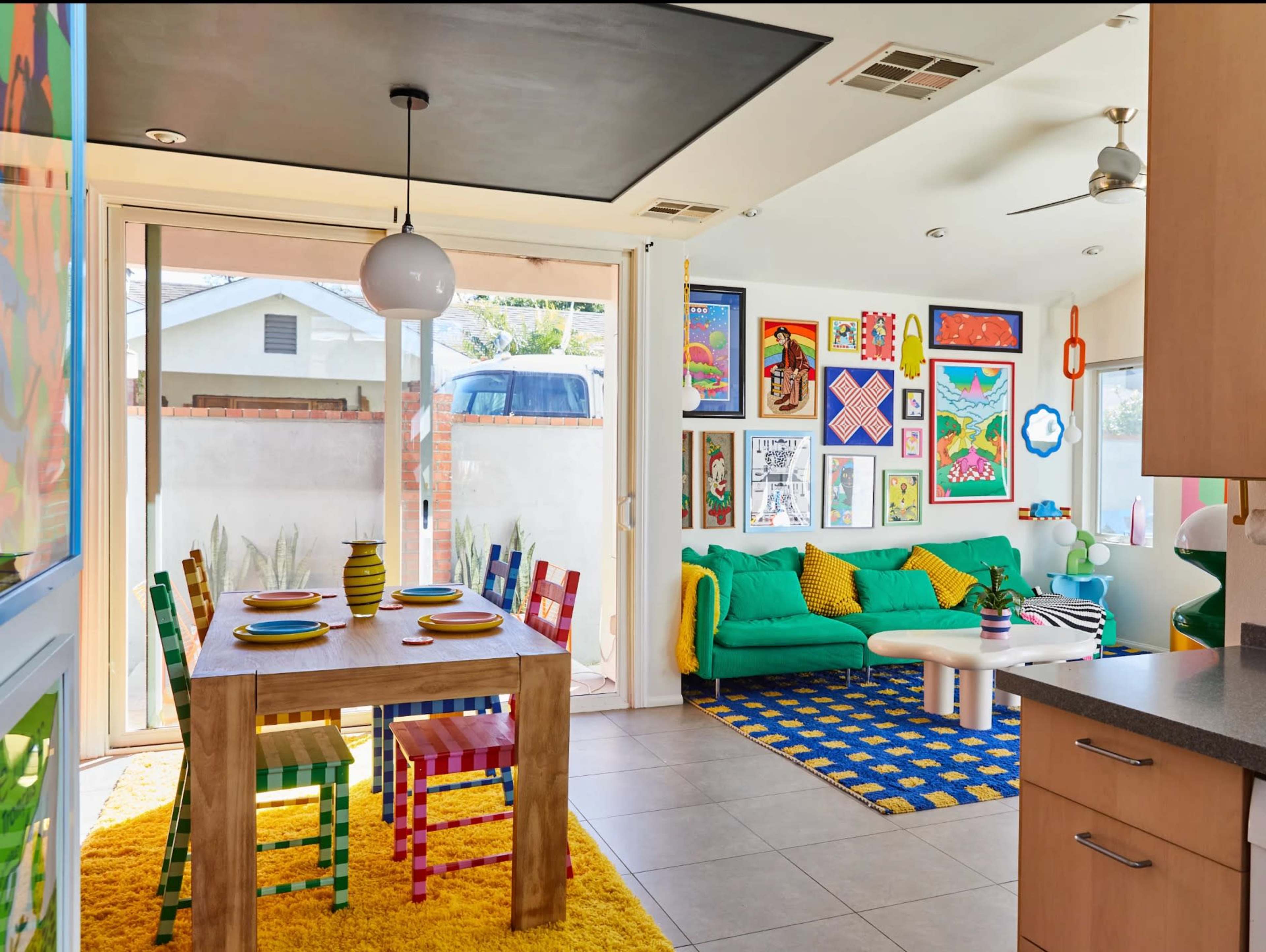 The image depicts a brightly colored living space featuring a dining area with a wooden table and multicolored chairs, alongside a cozy seating area adorned with vibrant art and decorative cushions.