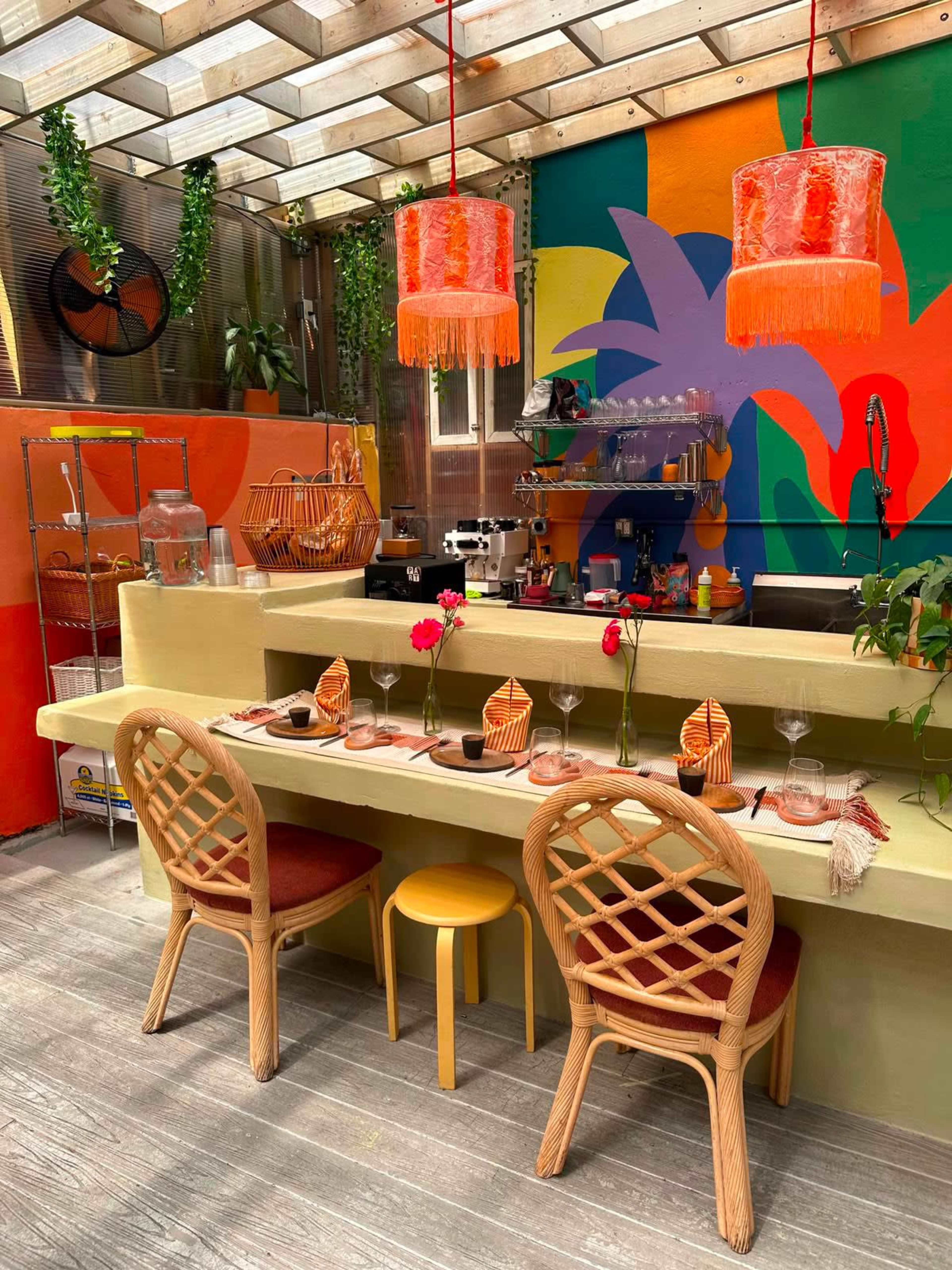 The image shows a colorful cafe interior with a vibrant mural, a long table set for dining, wicker chairs, and pendant lighting.