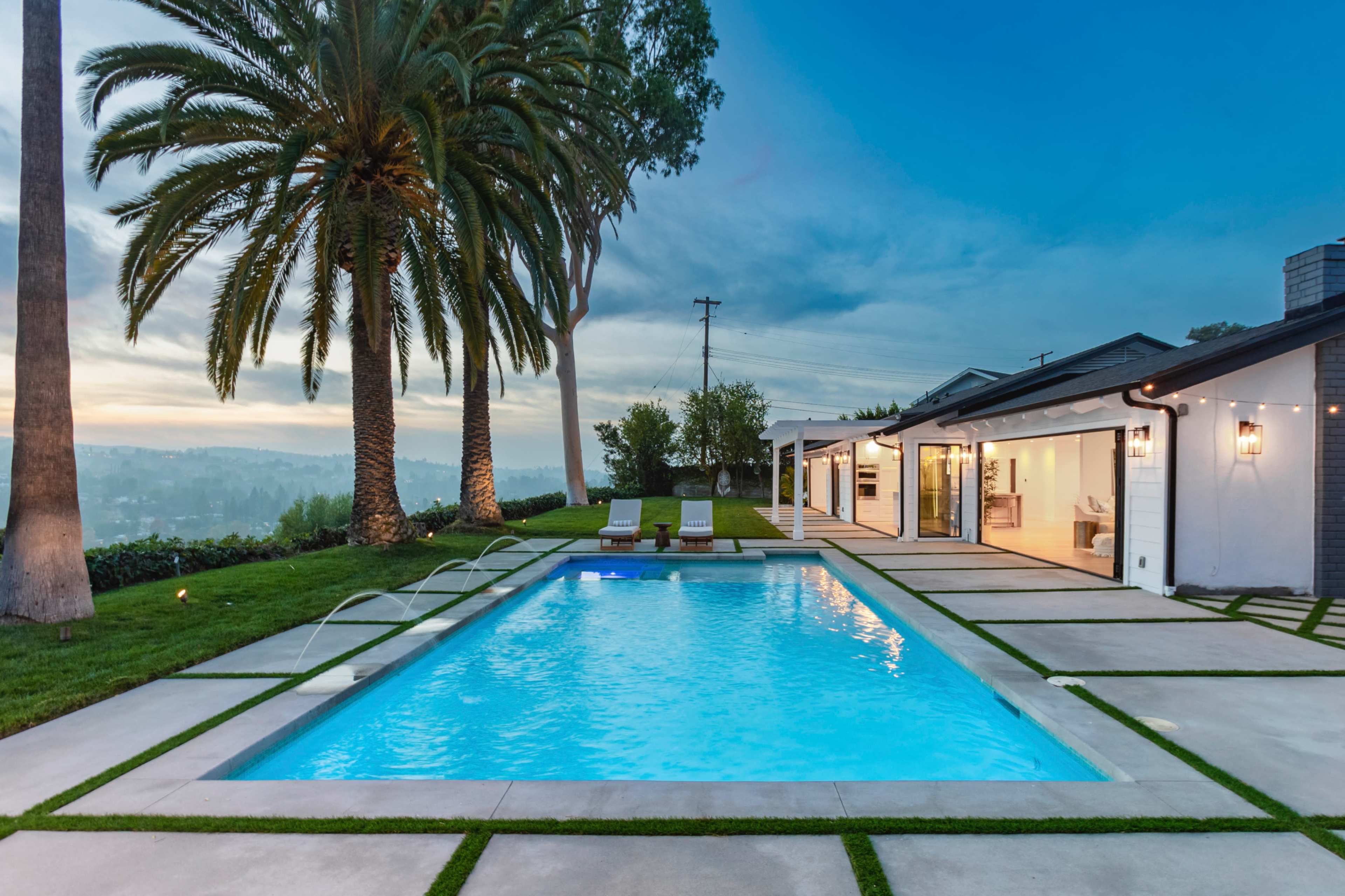 A modern home with a swimming pool surrounded by palm trees and well-maintained landscaping under a blue sky.