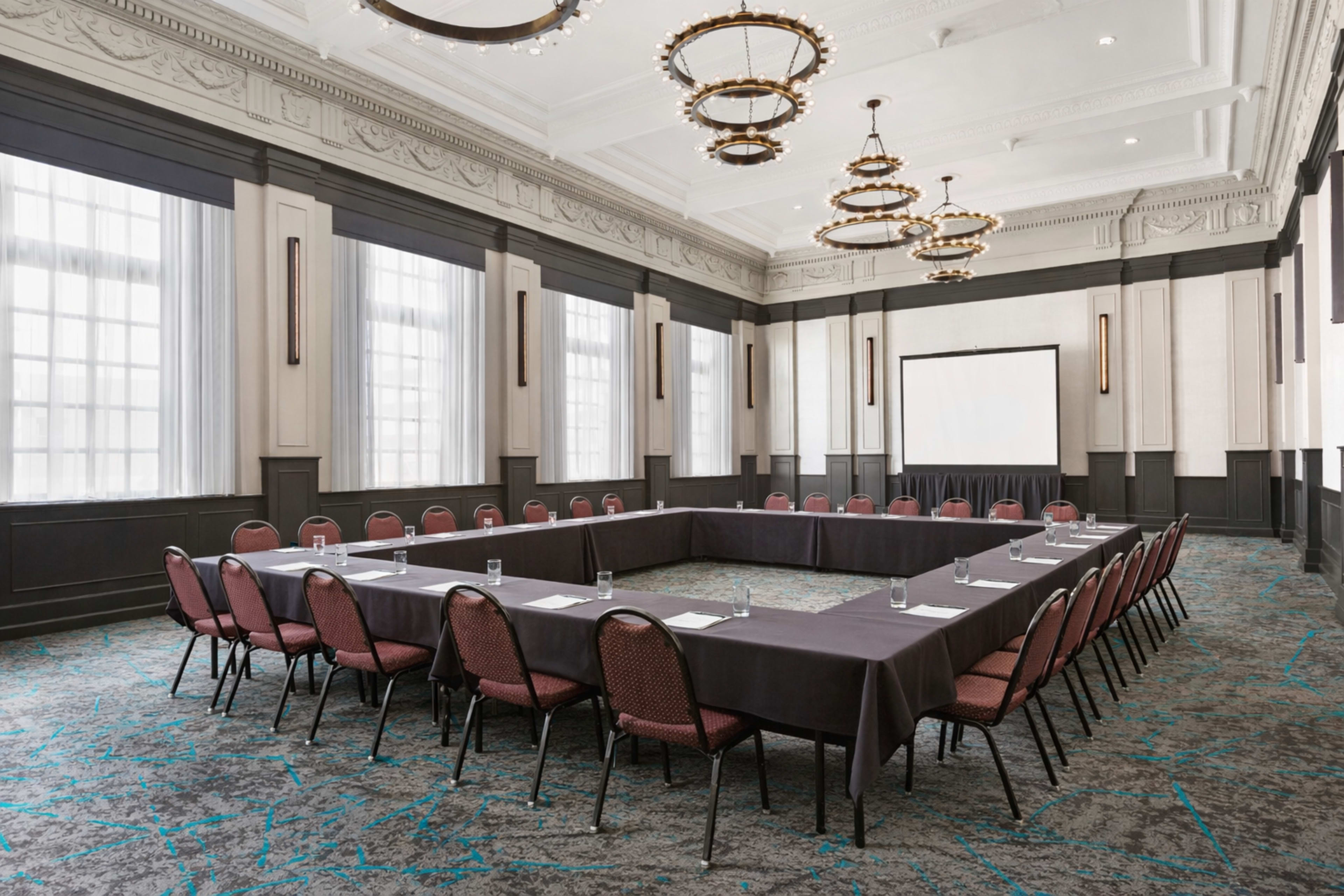 A large conference room features a rectangular arrangement of tables with chairs and a projection screen at one end.