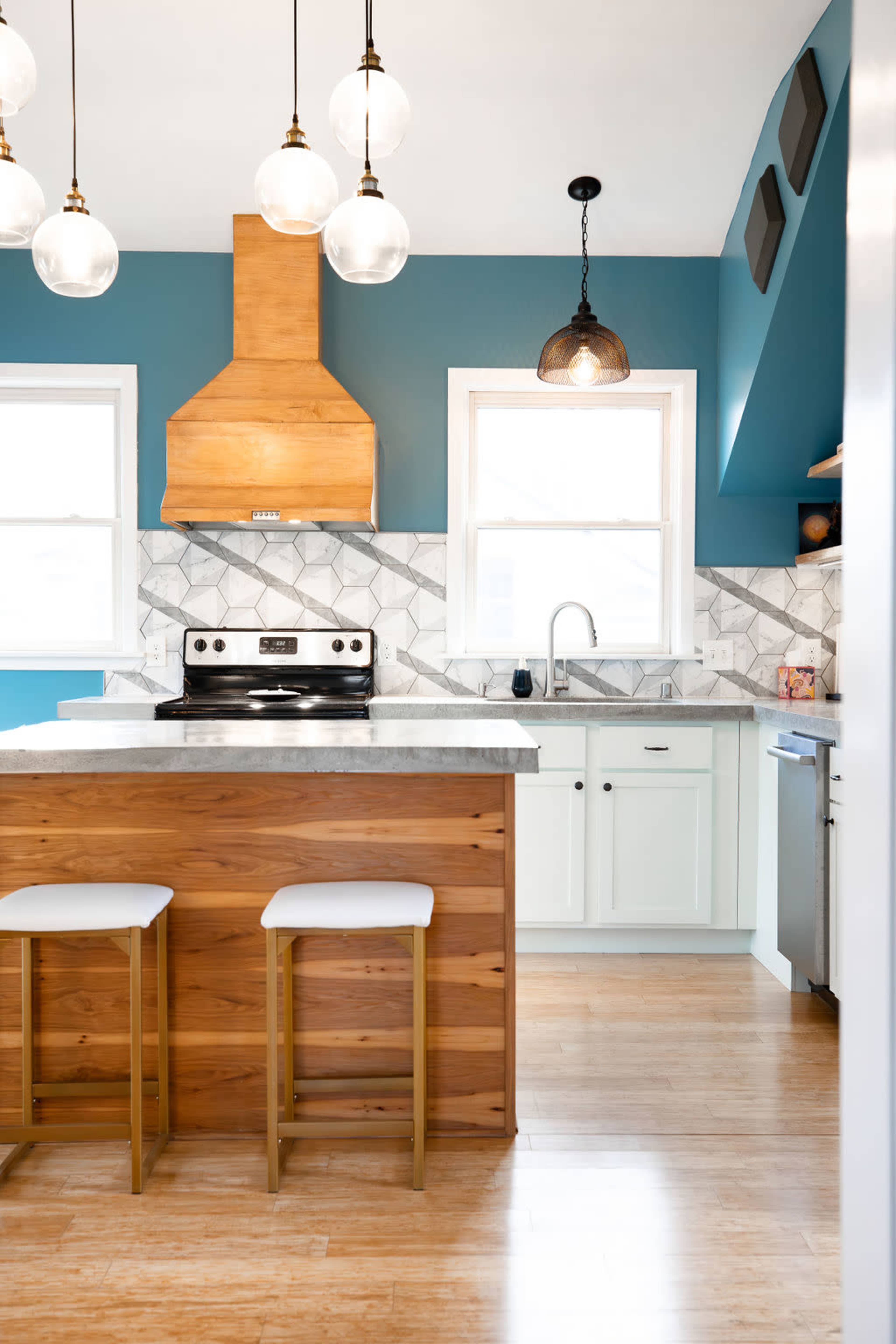 A modern kitchen features a wooden island with two white stools, a stainless steel stove, blue walls, and hanging pendant lights over the counter.