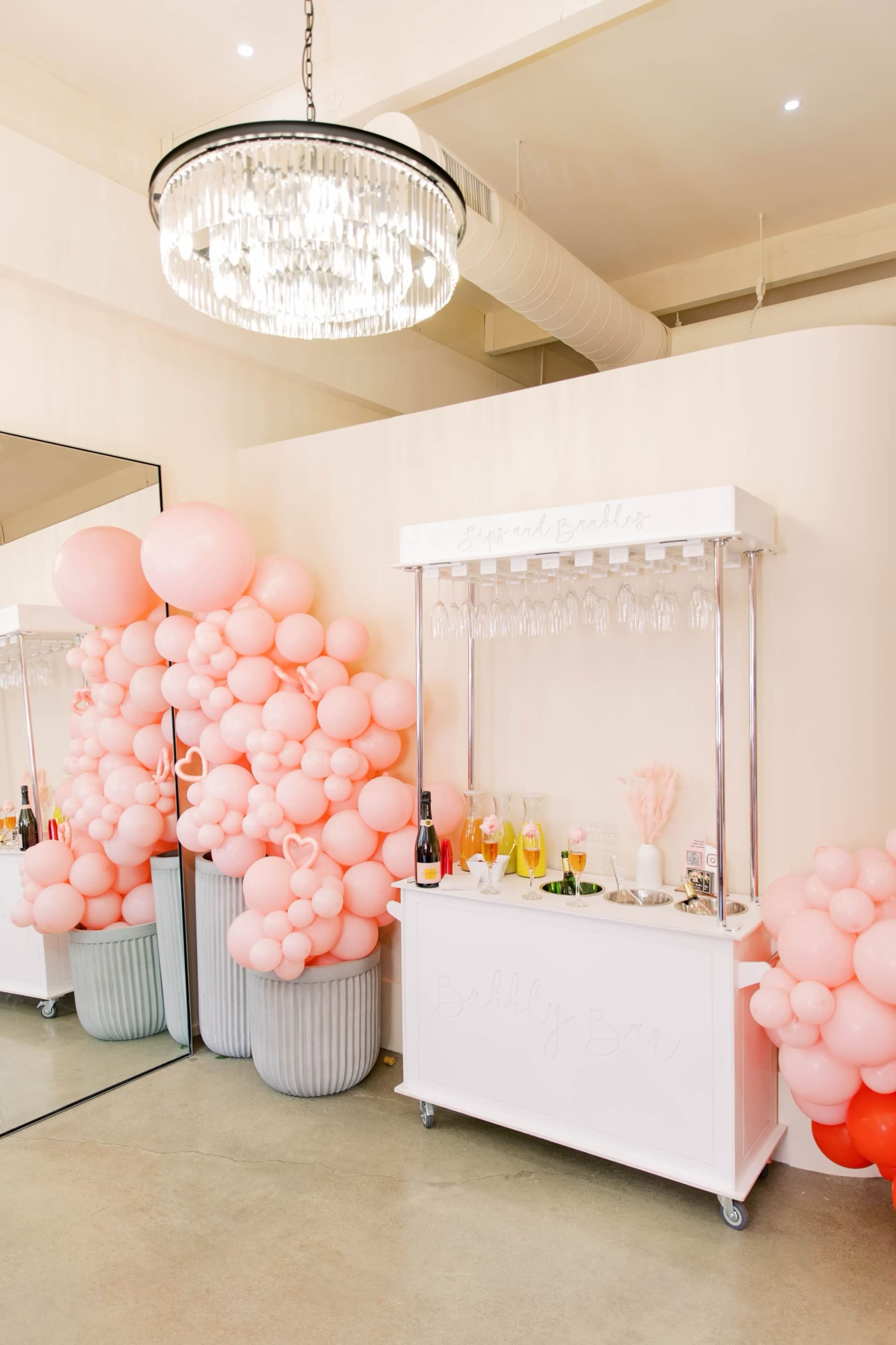 A white bar cart decorated with pastel pink balloons is set against a mirrored wall beneath a crystal chandelier.