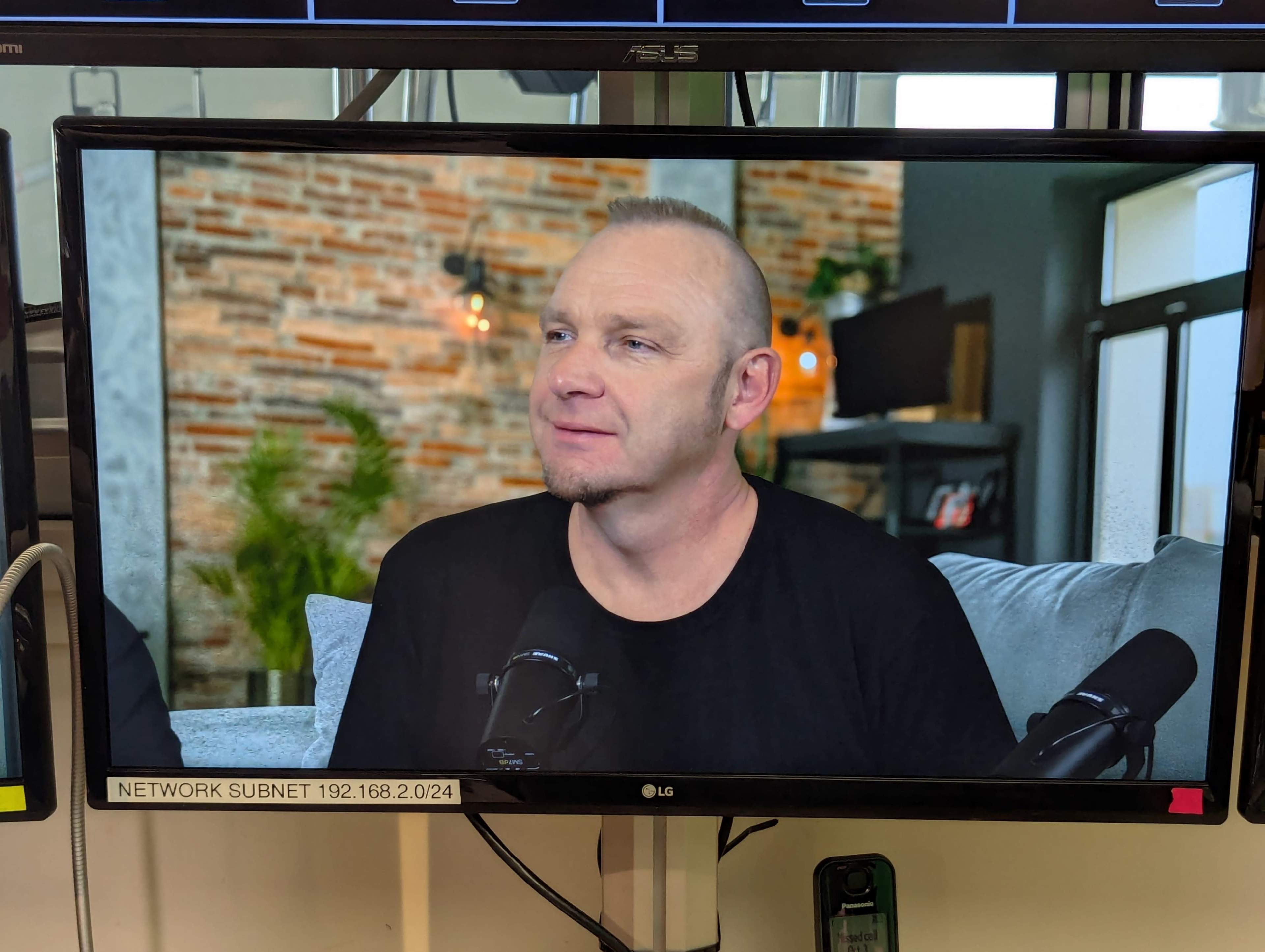 A man is seated on a couch in front of a microphone, with a brick wall and plants visible in the background on a television screen.