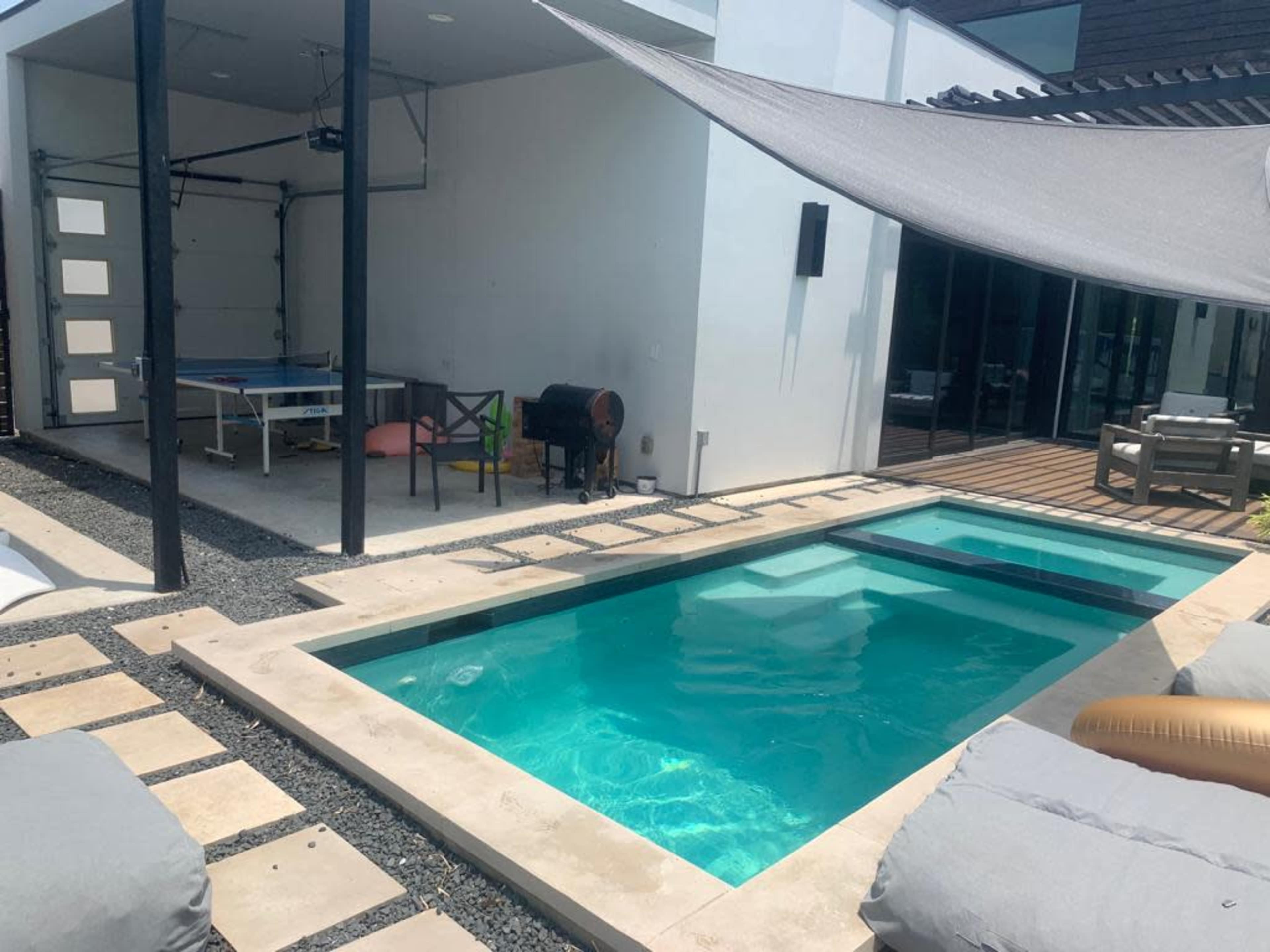 A modern patio area featuring a small swimming pool beside a covered garage with a table tennis setup and outdoor seating.