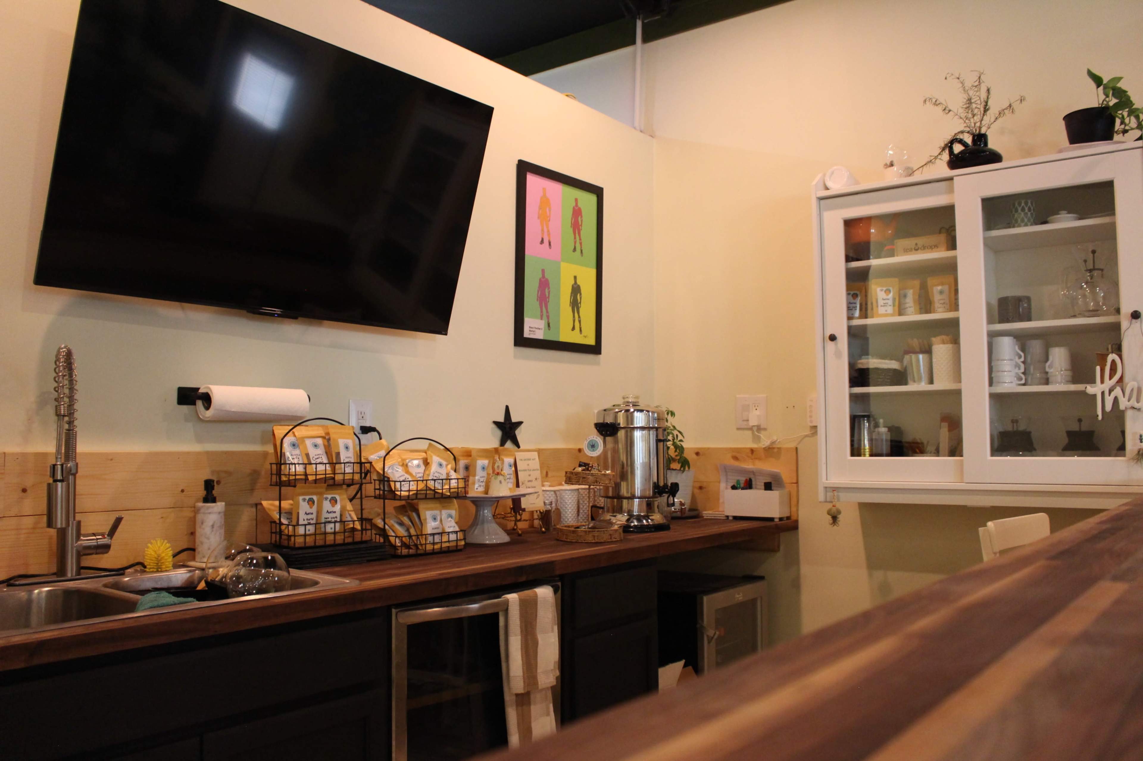 A kitchenette with a wall-mounted television, a sink, kitchen storage baskets, and a glass-front cabinet displaying various items.