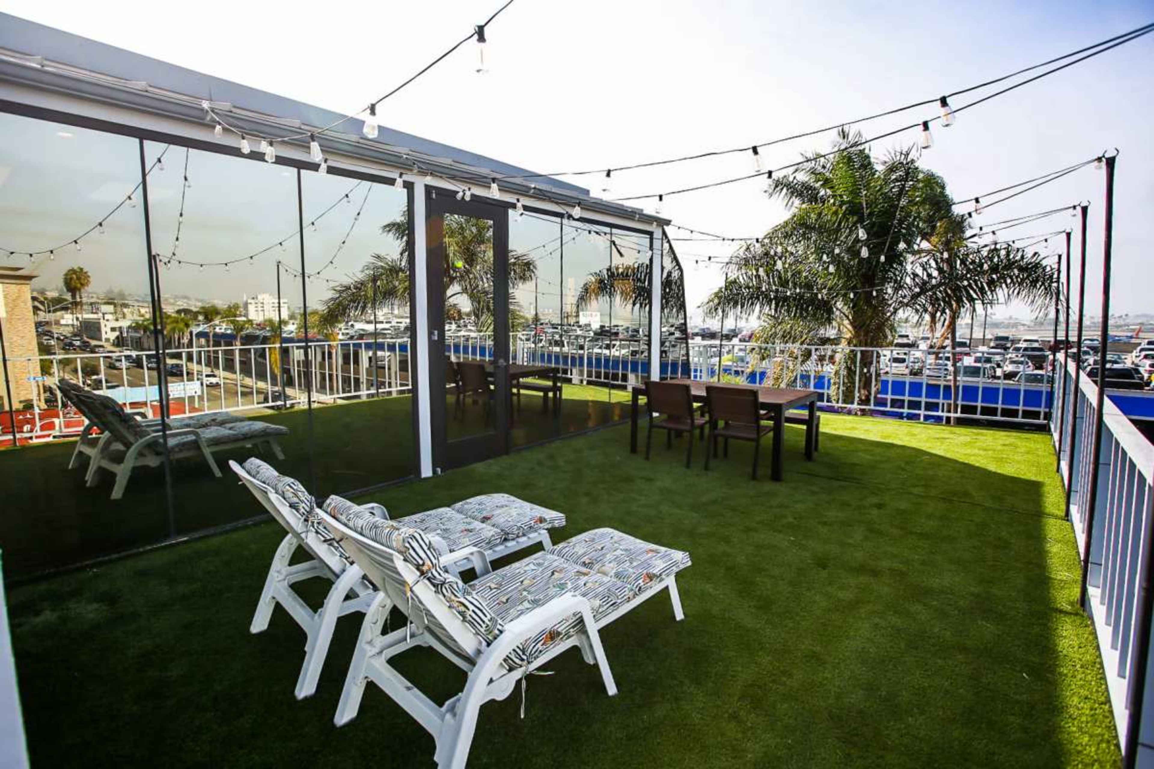 The image shows a rooftop area featuring two lounge chairs, a dining table with four chairs, and string lights overhead, surrounded by palm trees and a view of a parking lot.