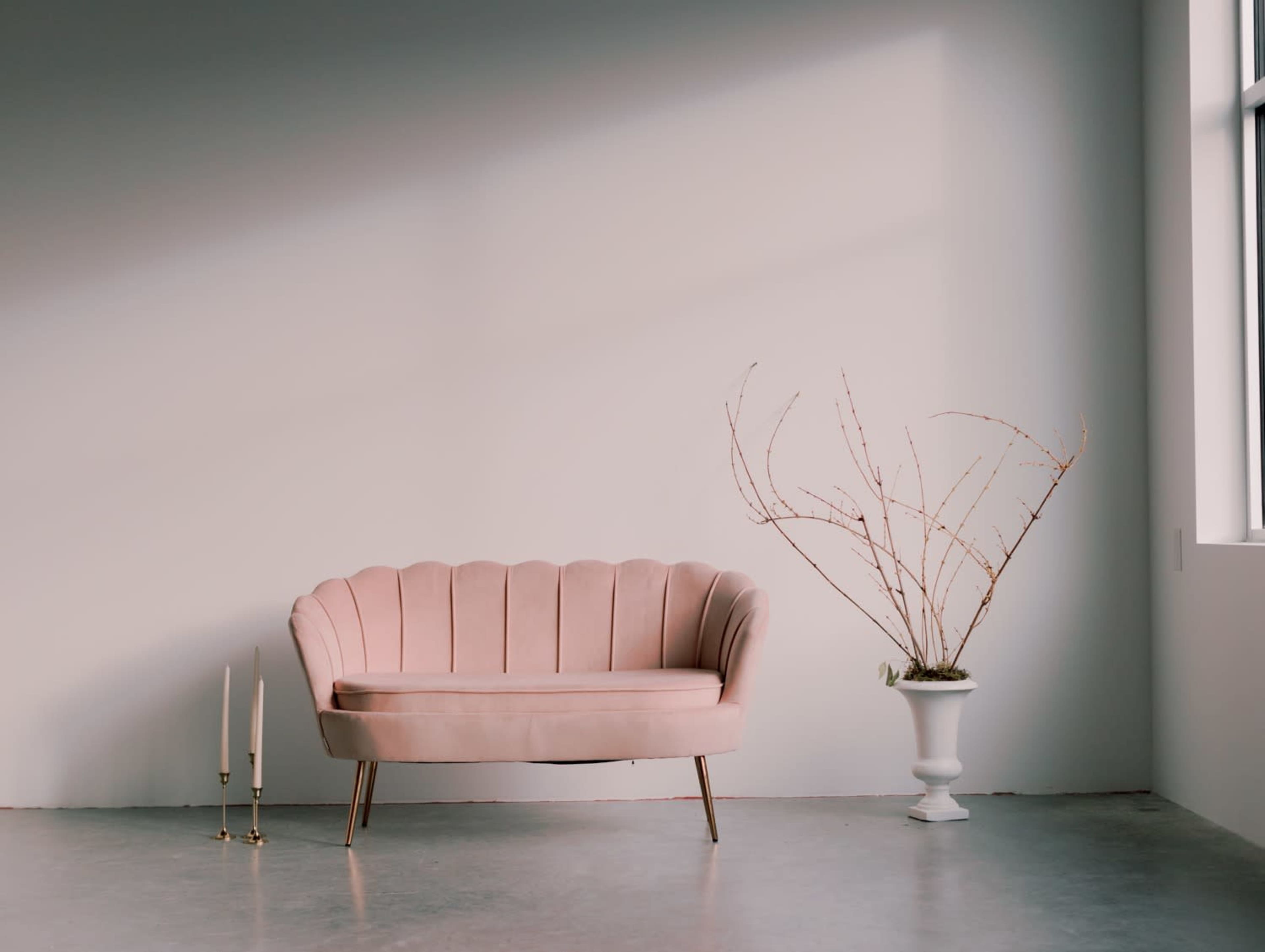 A pink scalloped sofa is placed against a light wall, accompanied by a slender vase with bare branches and two gold candle holders on the floor.