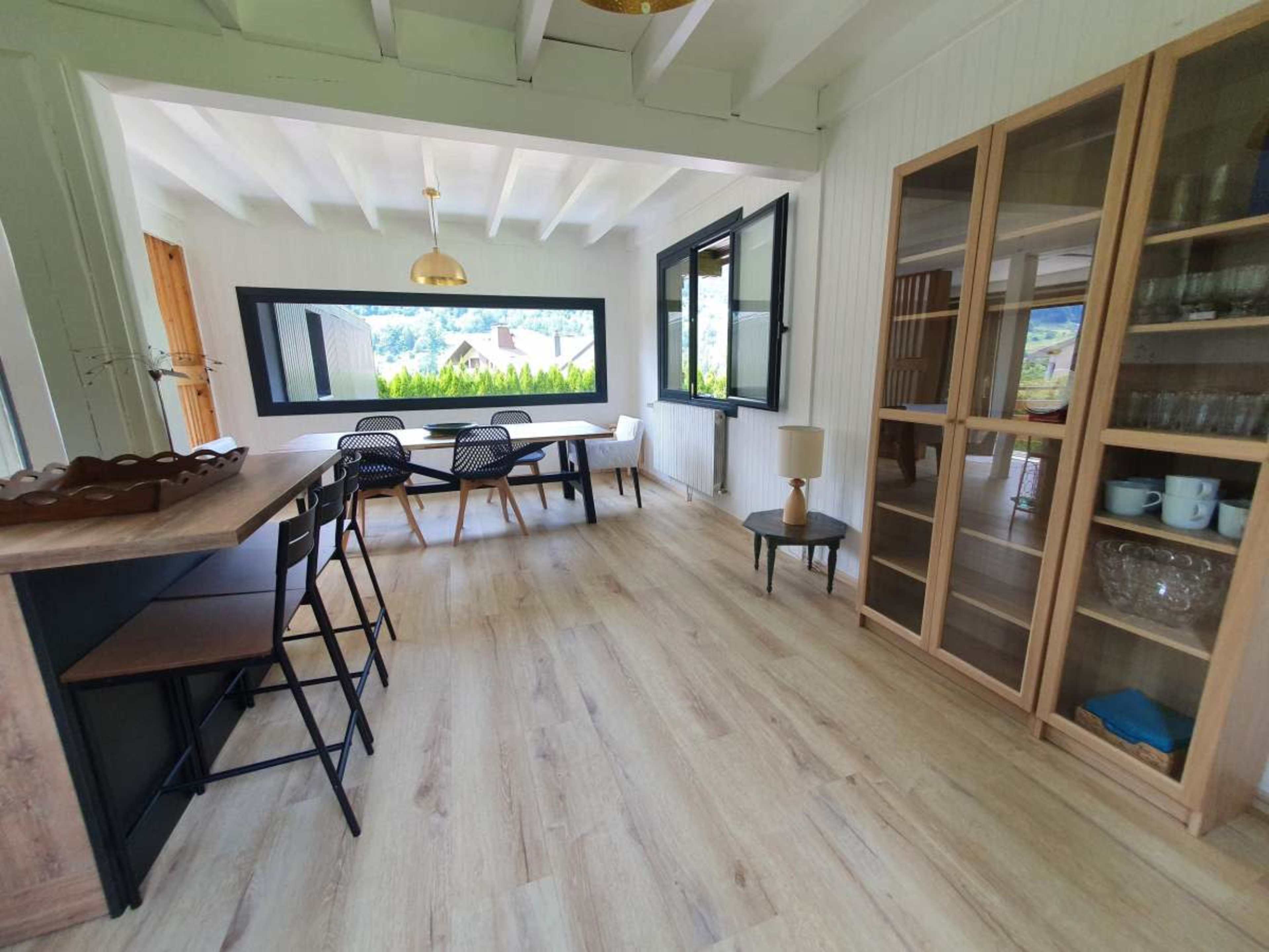 The image shows a bright, open dining area featuring a wooden table, a set of chairs, and a cabinet with glassware.