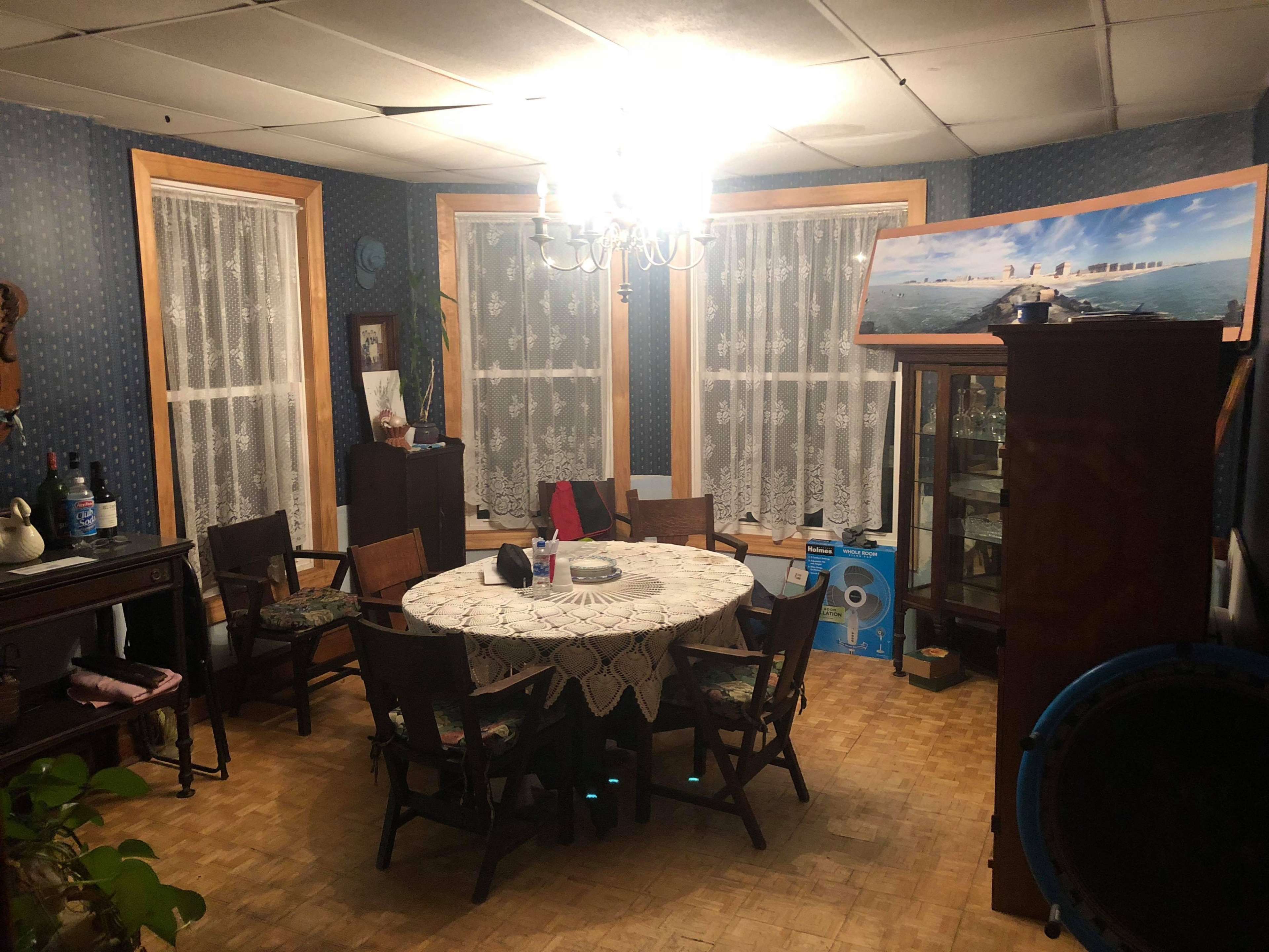 The image shows a dining room with a round table surrounded by wooden chairs, lace curtains on the windows, a chandelier overhead, and a cabinet displaying various items.