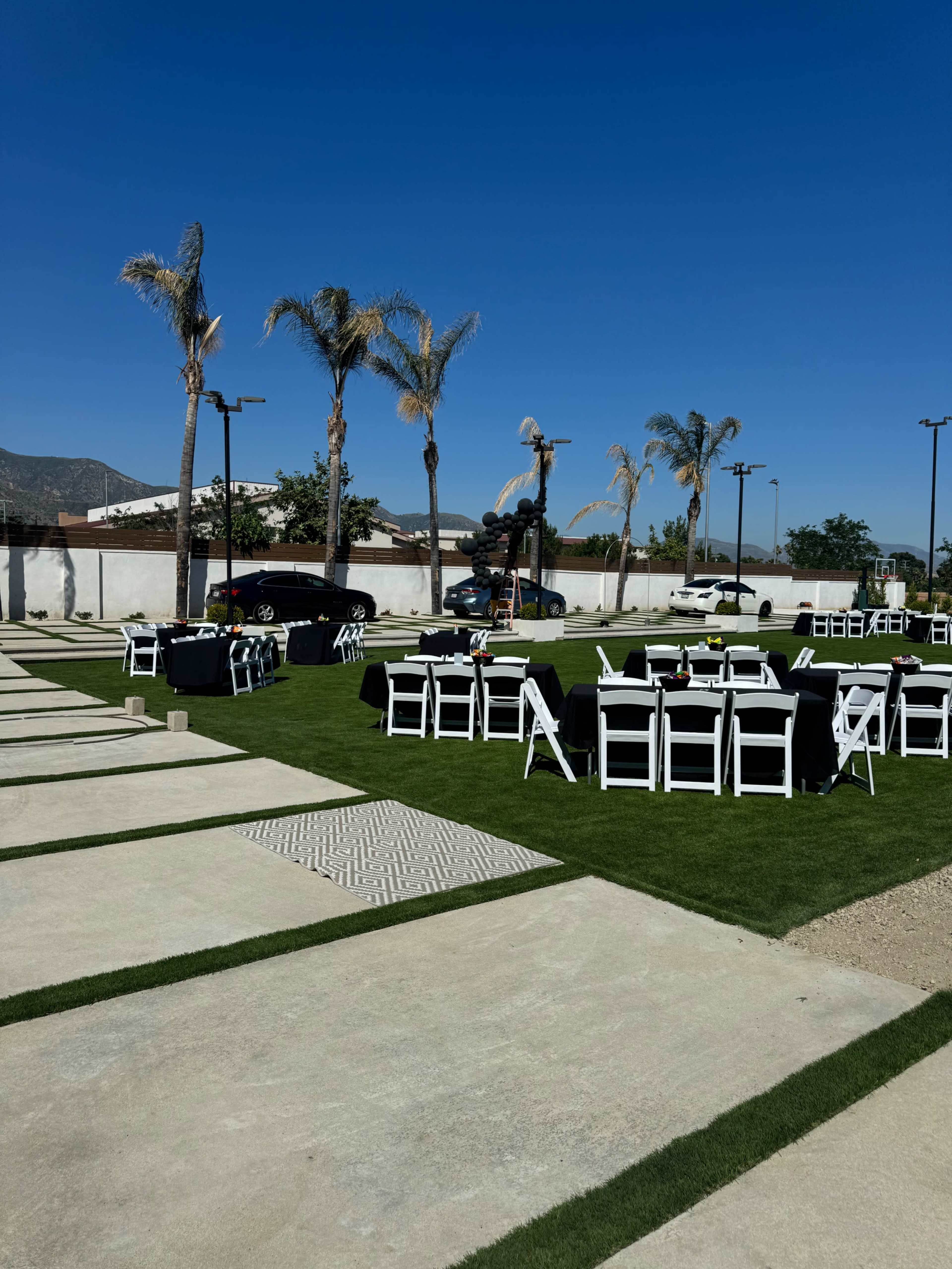 The scene features a landscaped outdoor area set with black and white tables and chairs, surrounded by palm trees and a clear blue sky.