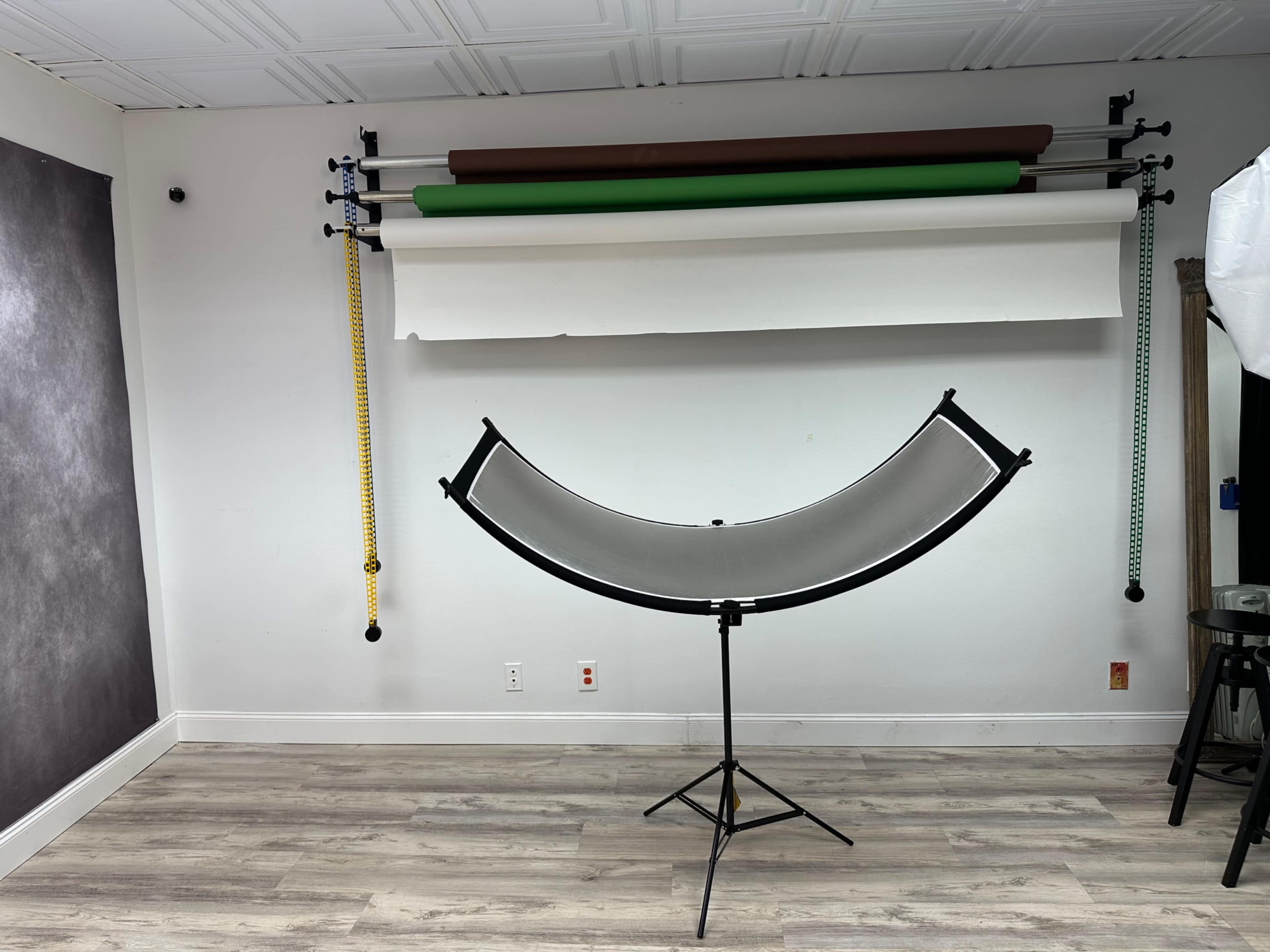 The image shows a studio setup with a curved backdrop support stand in front of a wall featuring several rolls of backdrop paper in various colors.