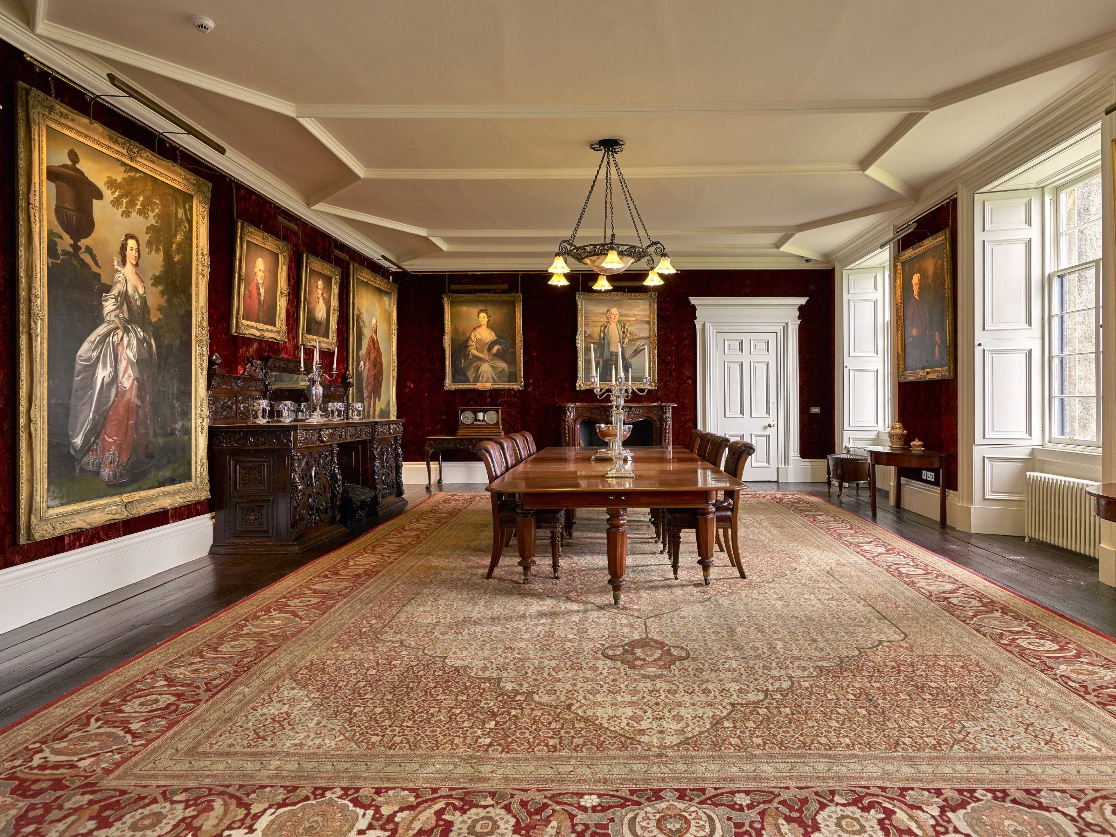 The image depicts a spacious, elegantly furnished dining room featuring a large wooden table, ornate ceiling lights, and several framed portraits on the walls.