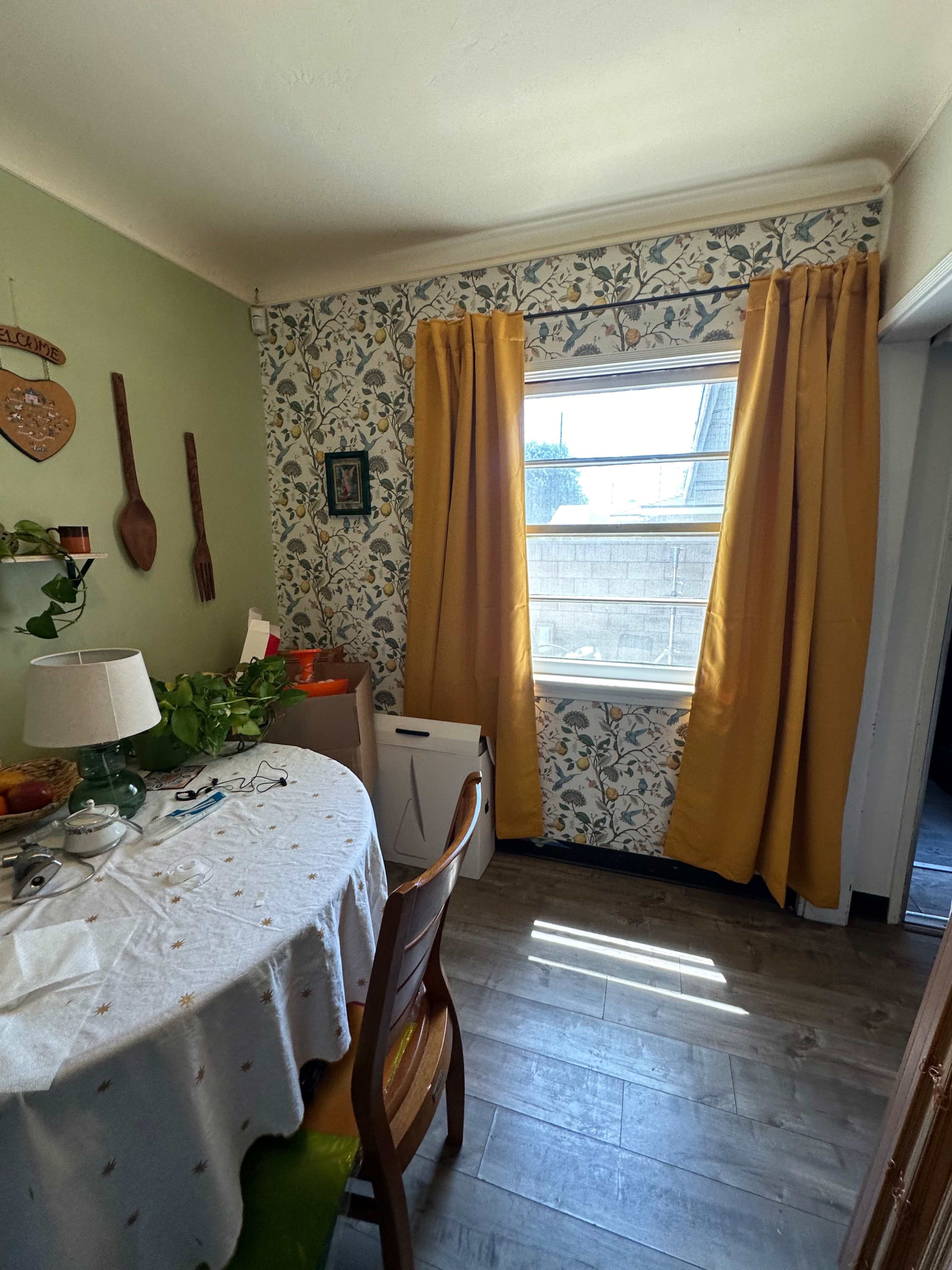 A small dining area features a round table covered with a tablecloth, surrounded by wooden chairs, and a window with yellow curtains that overlooks an outdoor space.