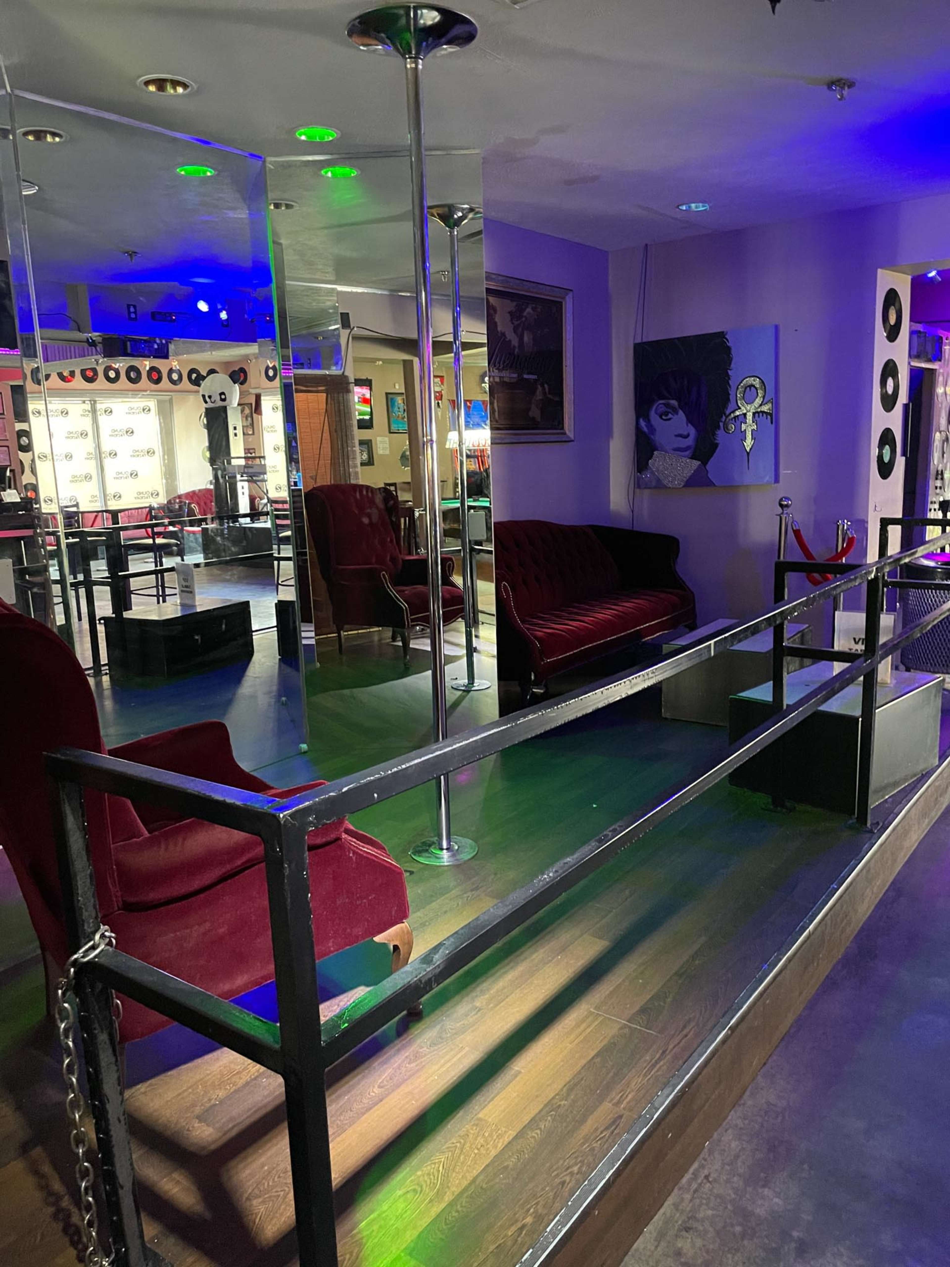 A small lounge area with two red velvet chairs and a mirrored wall, illuminated by colorful lights, alongside a stage area featuring a microphone stand and a pole.