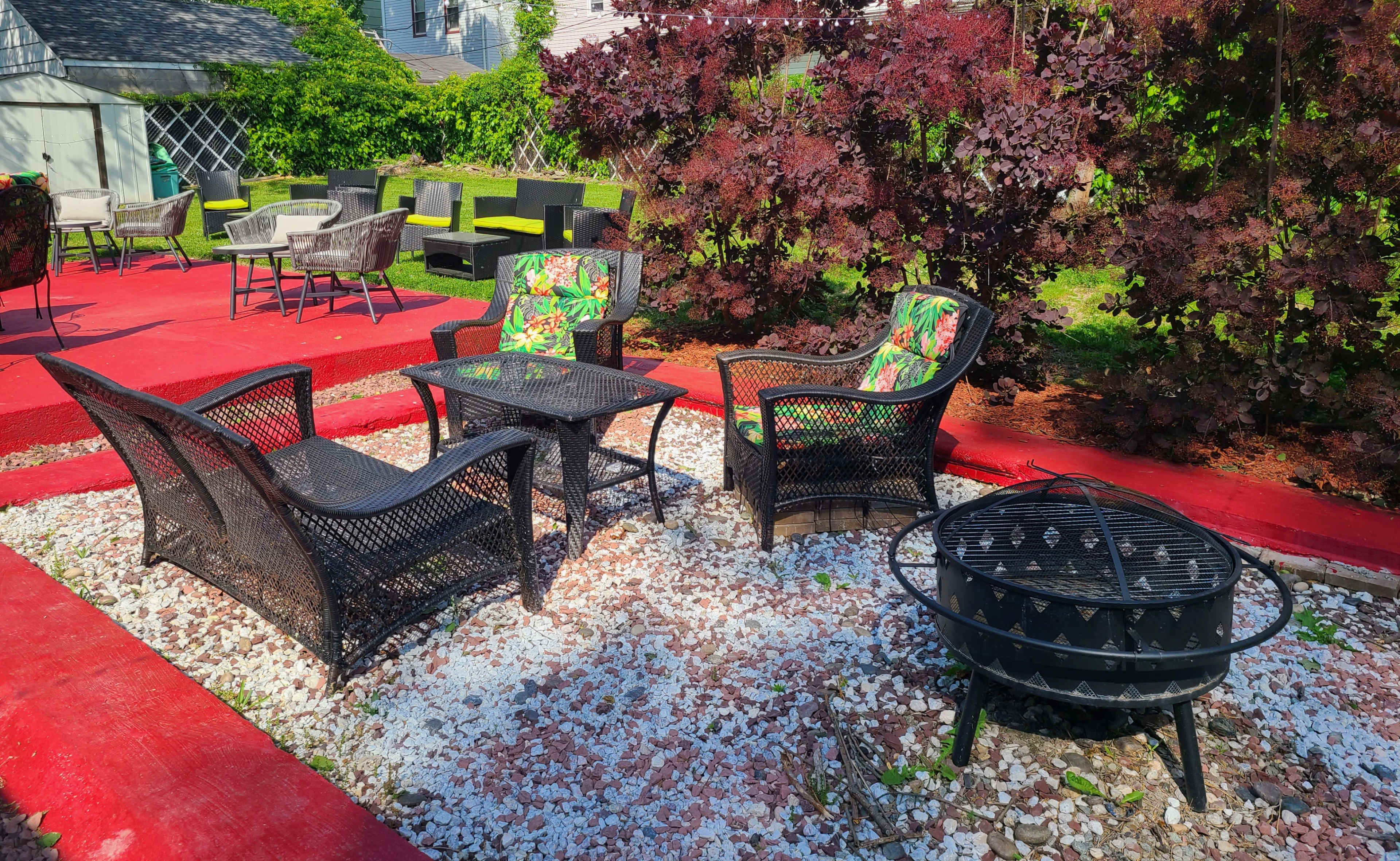 A patio area with four black wicker chairs and a small table arranged around a fire pit, surrounded by gravel and vibrant red landscaping.
